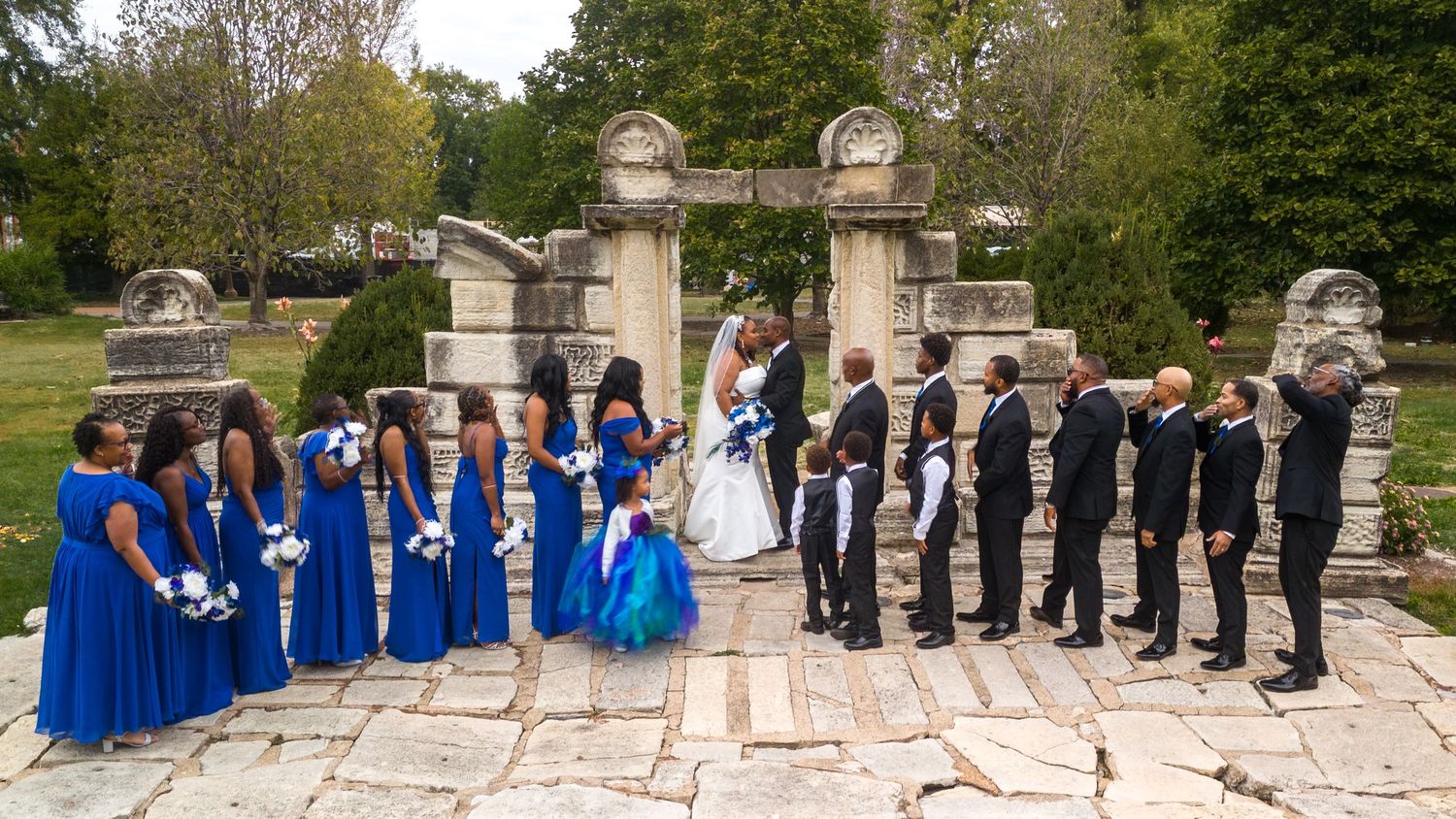 Wedding party in blue dresses and black suits poses symmetrically on stone steps outdoors.