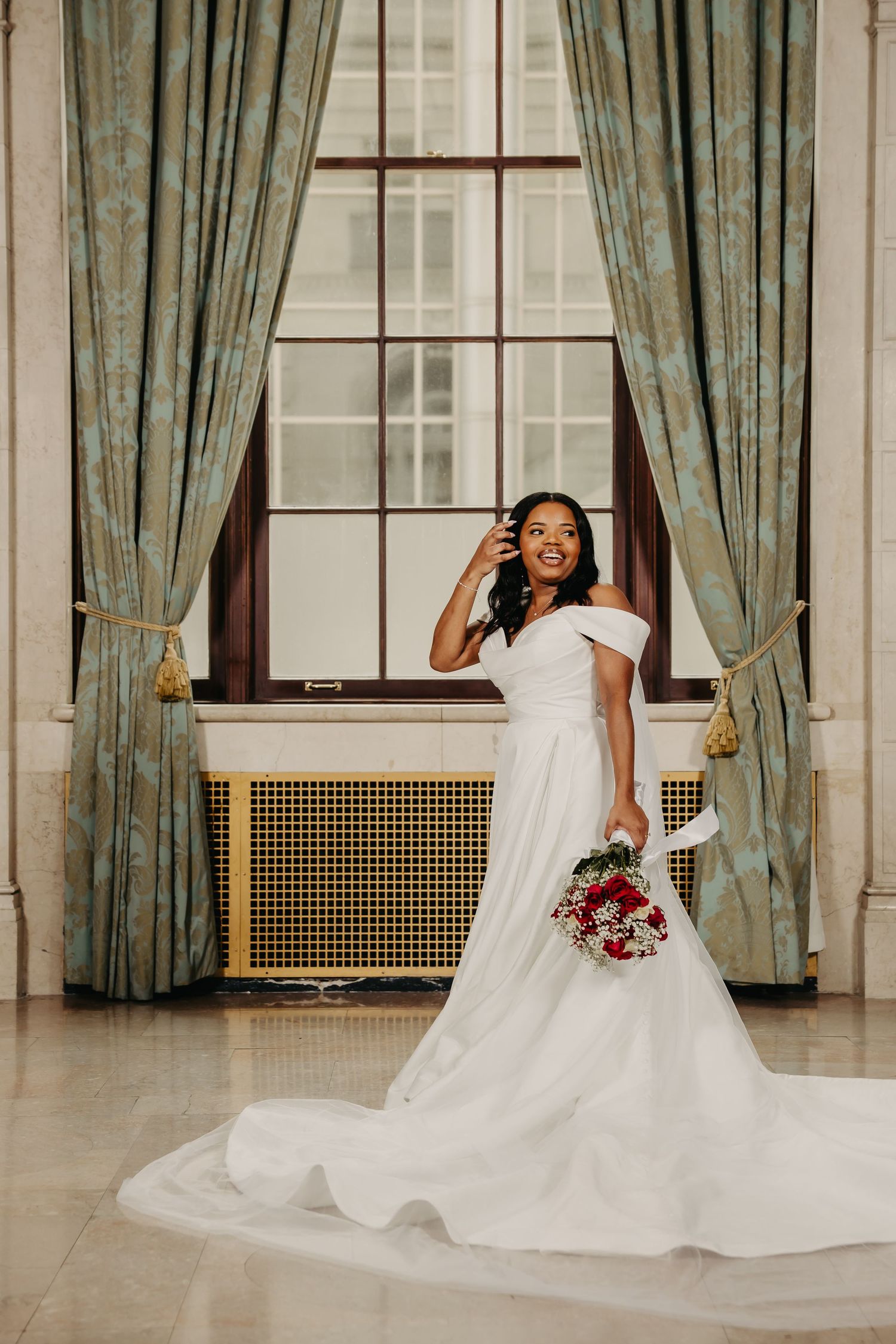 Bride in flowing white gown poses with red bouquet against elegant curtained backdrop.