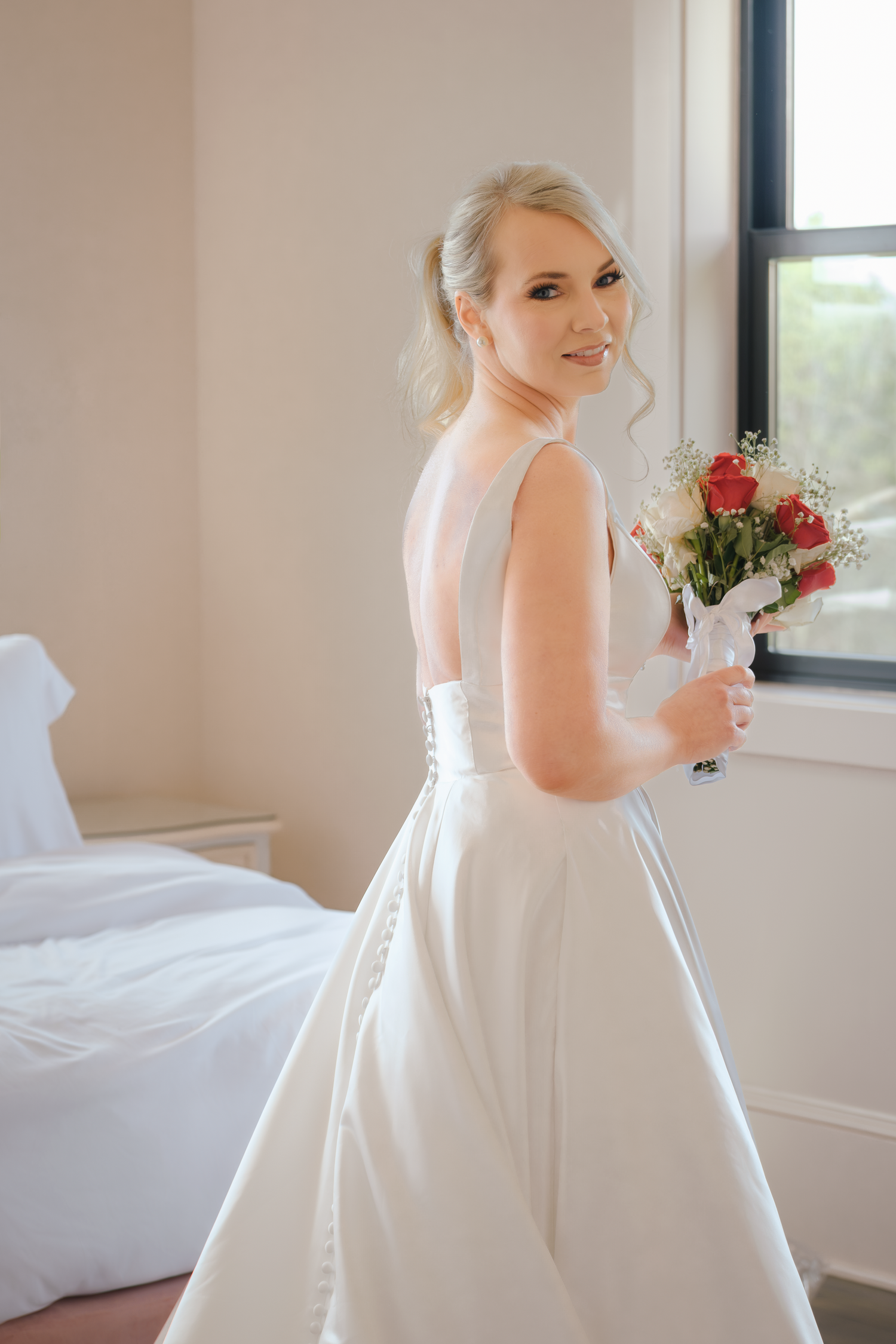 Blonde bride in white gown holding red rose bouquet, smiling over shoulder near window in bright room.