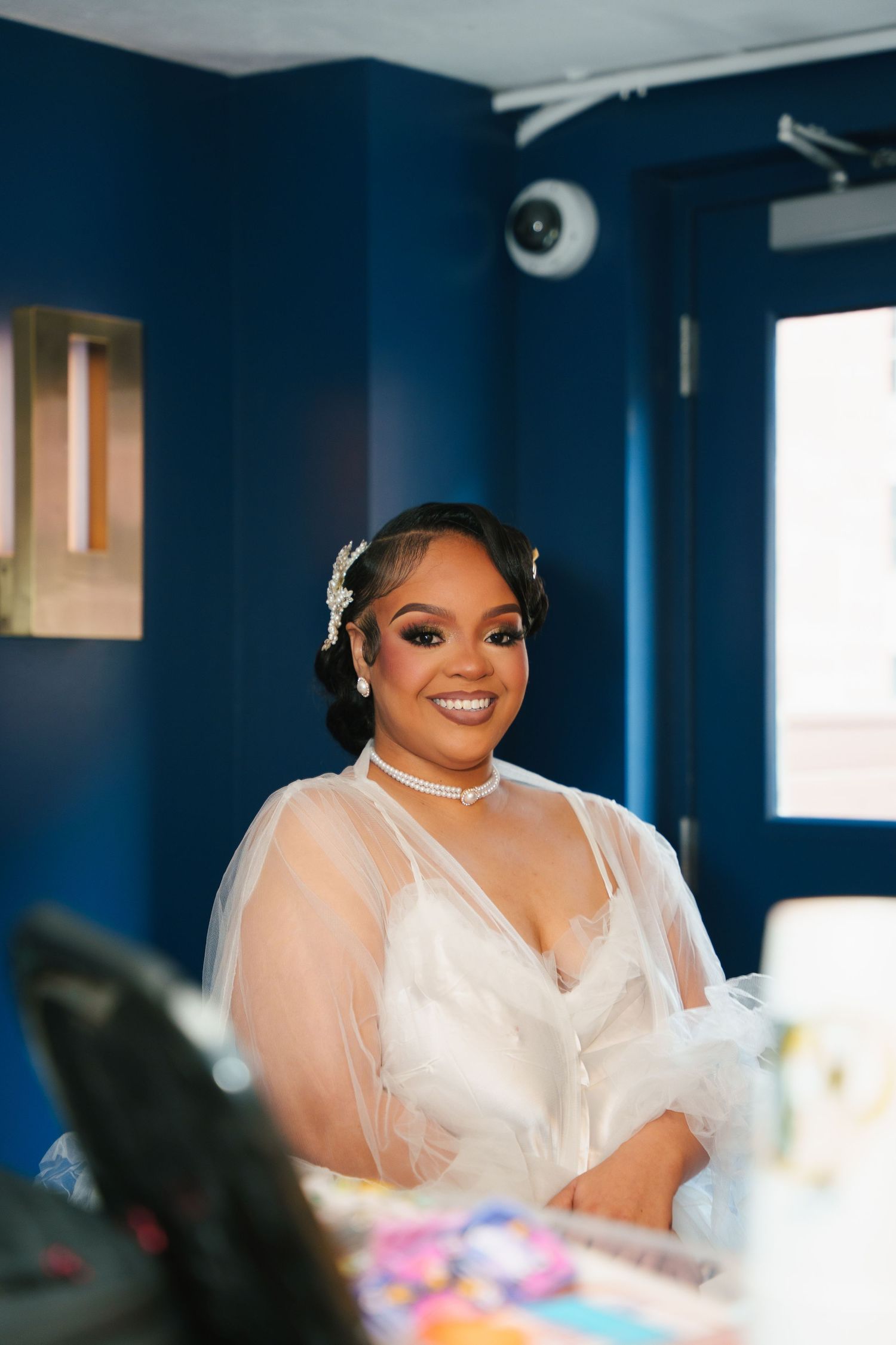 A bride in a white wedding dress and veil smiles radiantly while getting ready in a room with deep blue walls.