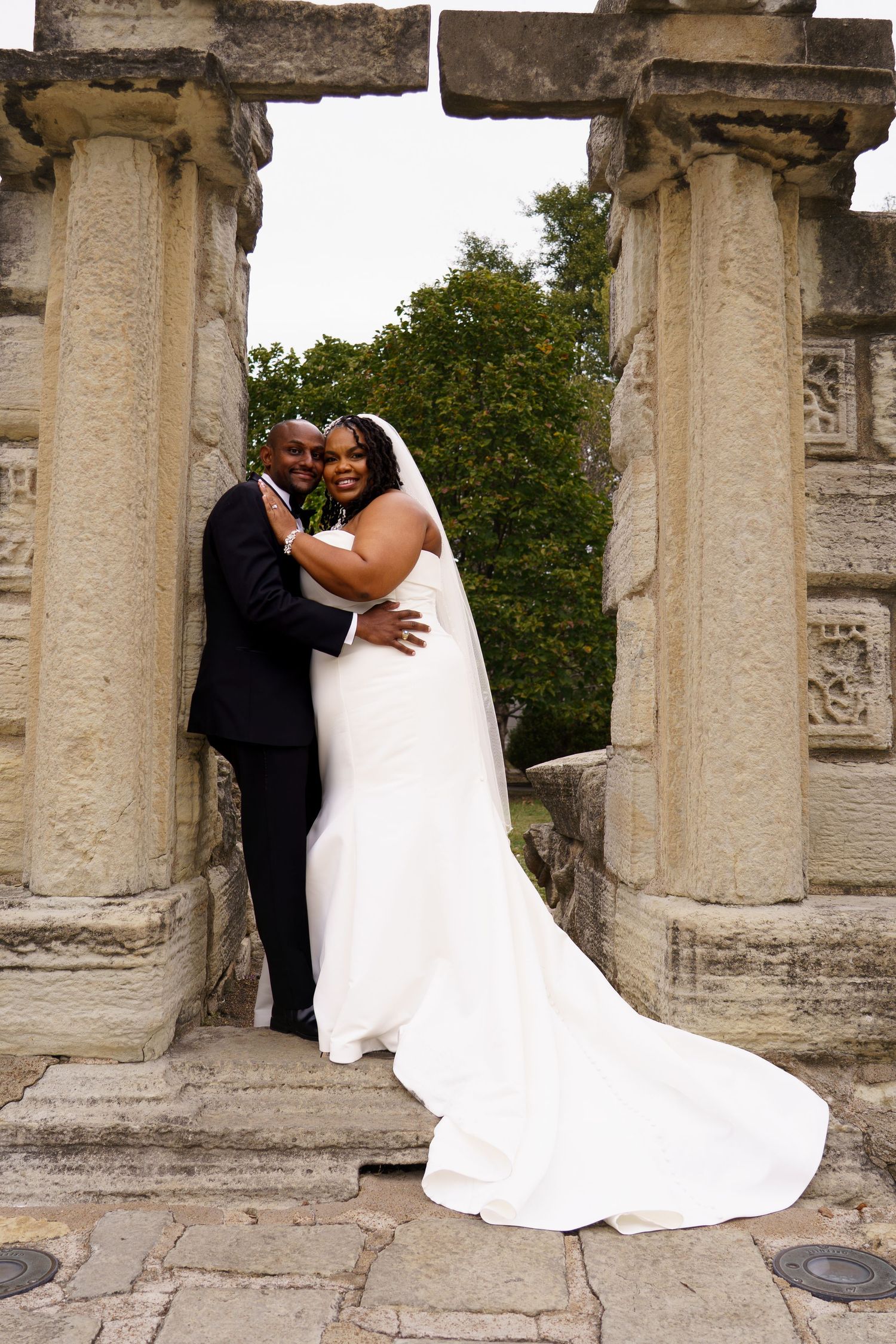 A couple in wedding attire embraces among ancient stone pillars and columns during an outdoor photoshoot.