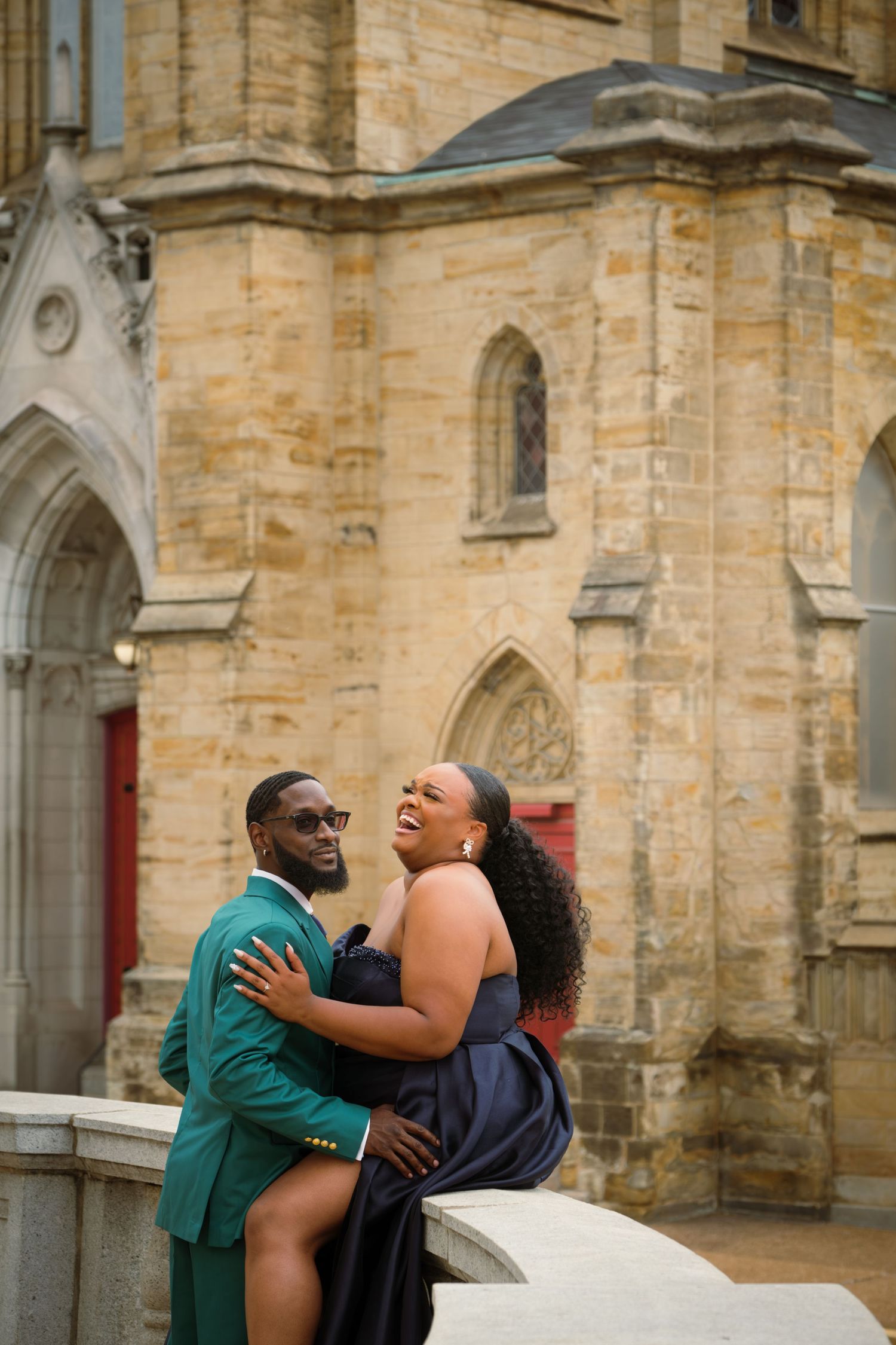 Elegant couple posing in front of a historic stone church, man in green suit and woman in navy gown, laughing joyfully.