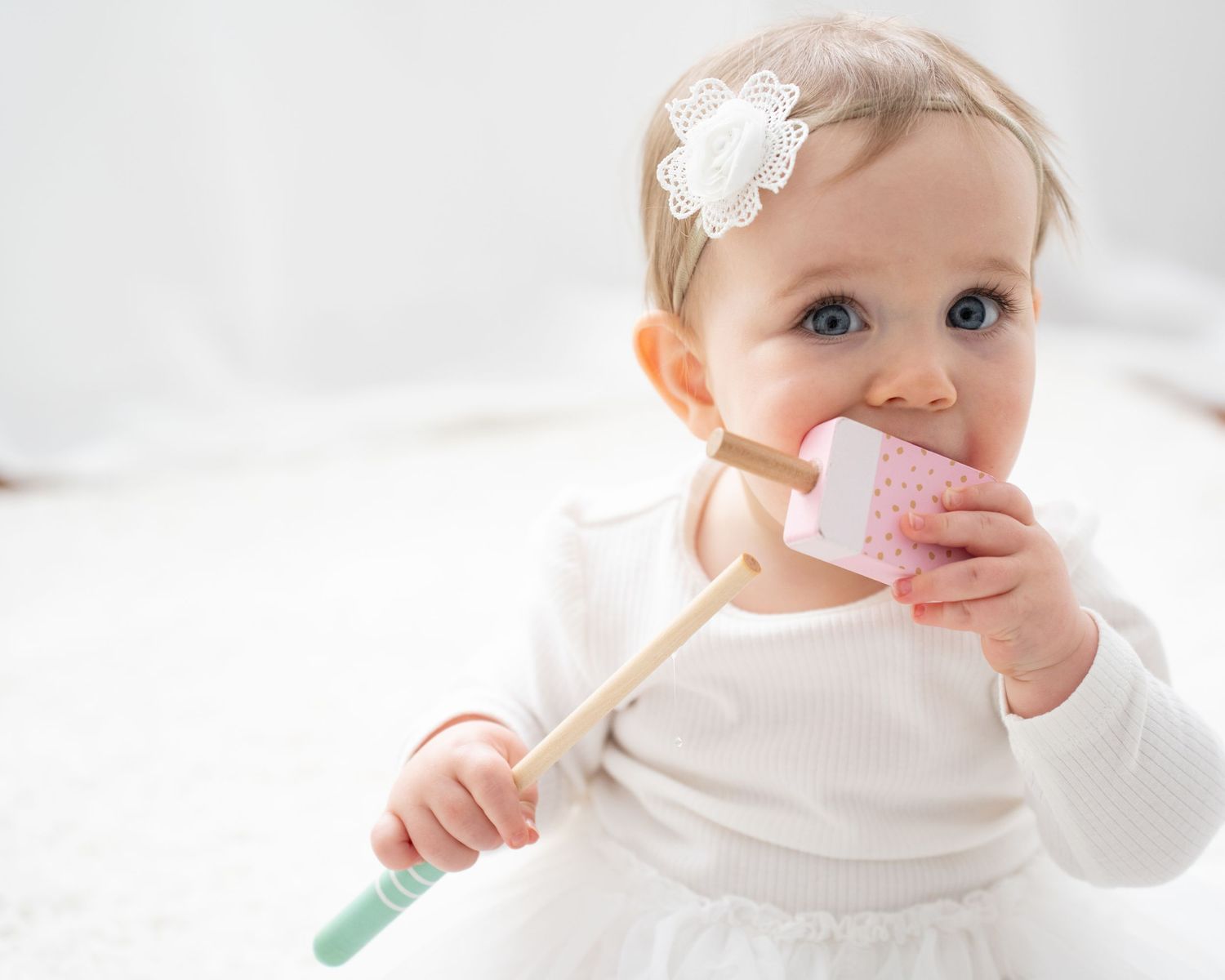 Adorable baby girl in white outfit chewing on a wooden toy ice cream while wearing a floral headband.