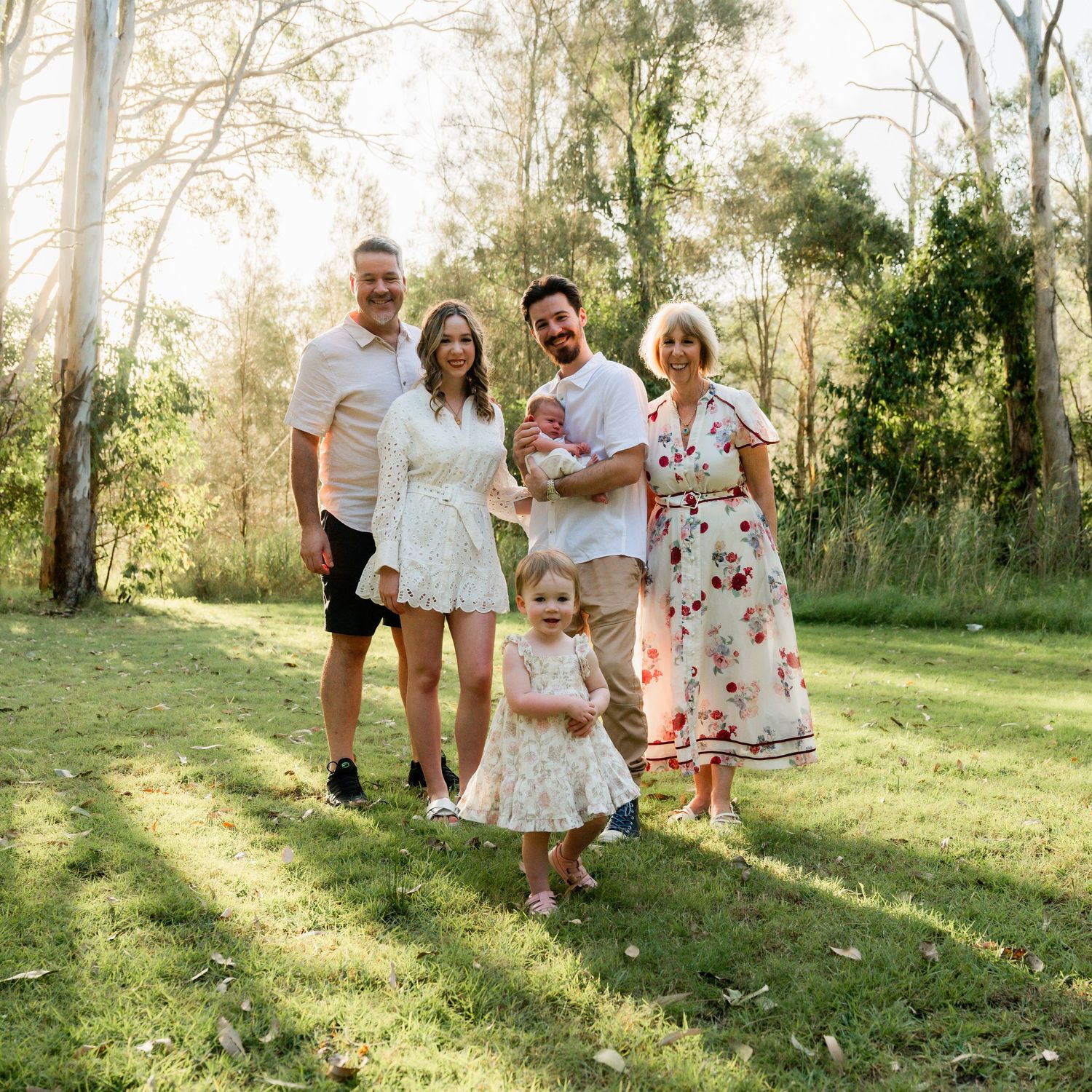 Family of five posing outdoors in a sunny park, wearing white and floral outfits, with tall trees in the background.