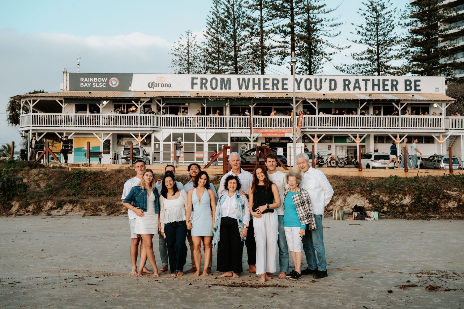 Family group photo on beach in front of Rainbow Bay Surf Club sign reading From Where You'd Rather Be.