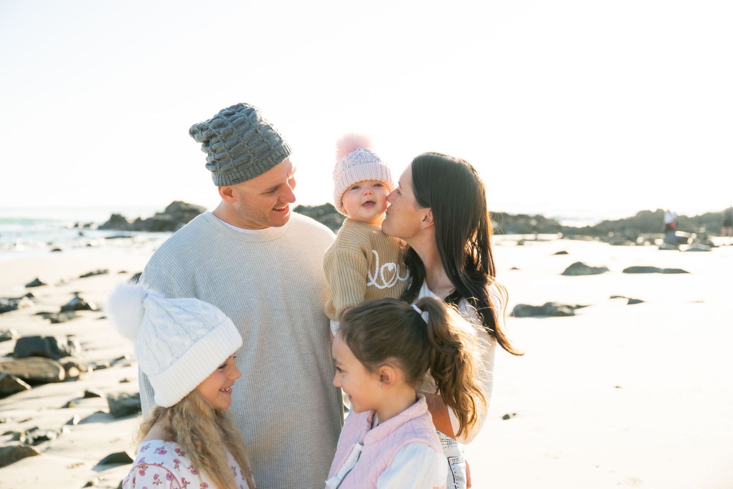Family of five enjoying a sunny beach day, parents kissing baby while two girls stand close, rocks and ocean in background.