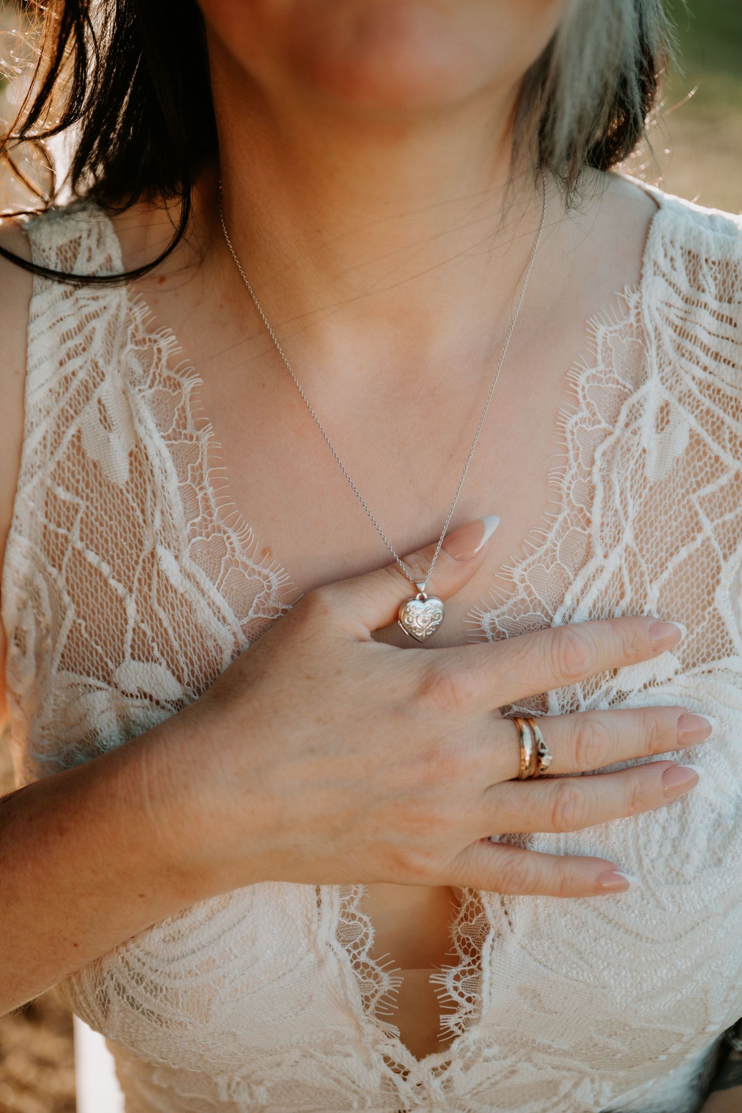 Bride in lace wedding dress holding heart locket necklace, wearing gold wedding band ring.