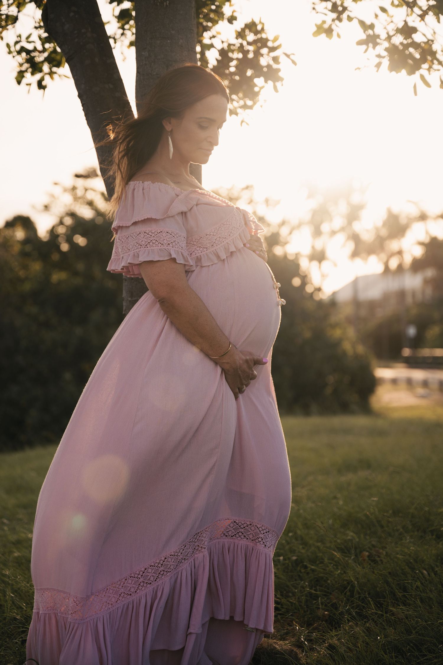 Pregnant woman in pink off-shoulder dress standing by tree at golden hour sunset in lush green park.