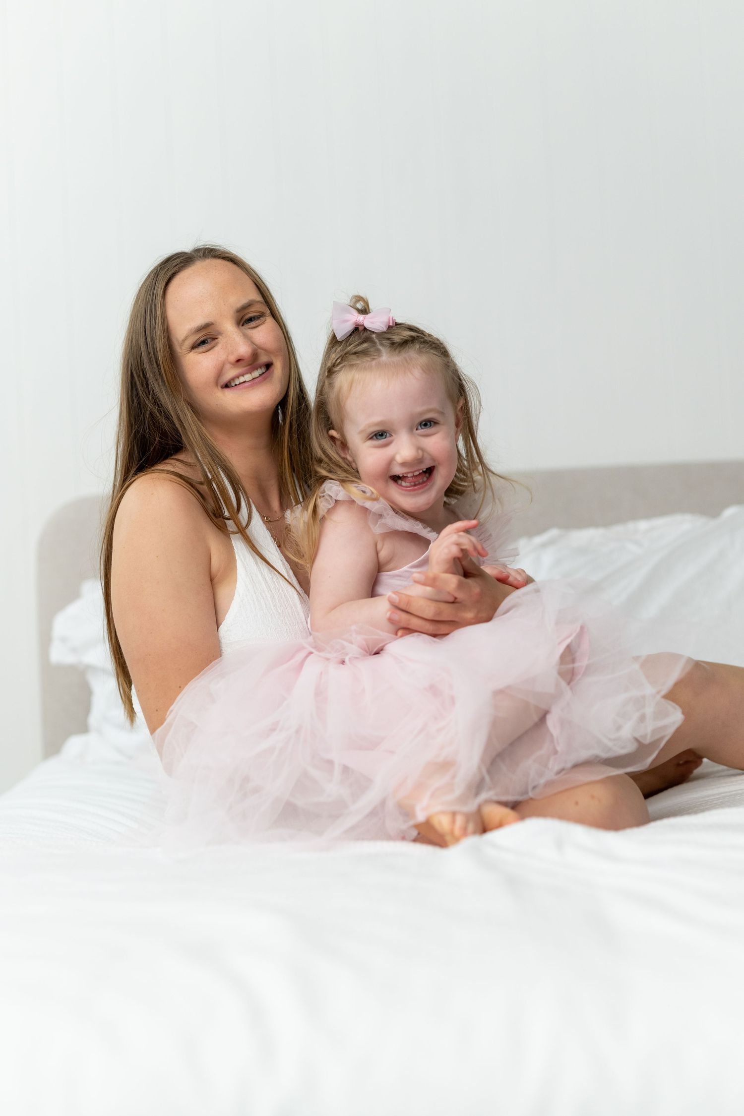 A smiling mother and her laughing toddler daughter in matching pink outfits sitting together on a white bed.