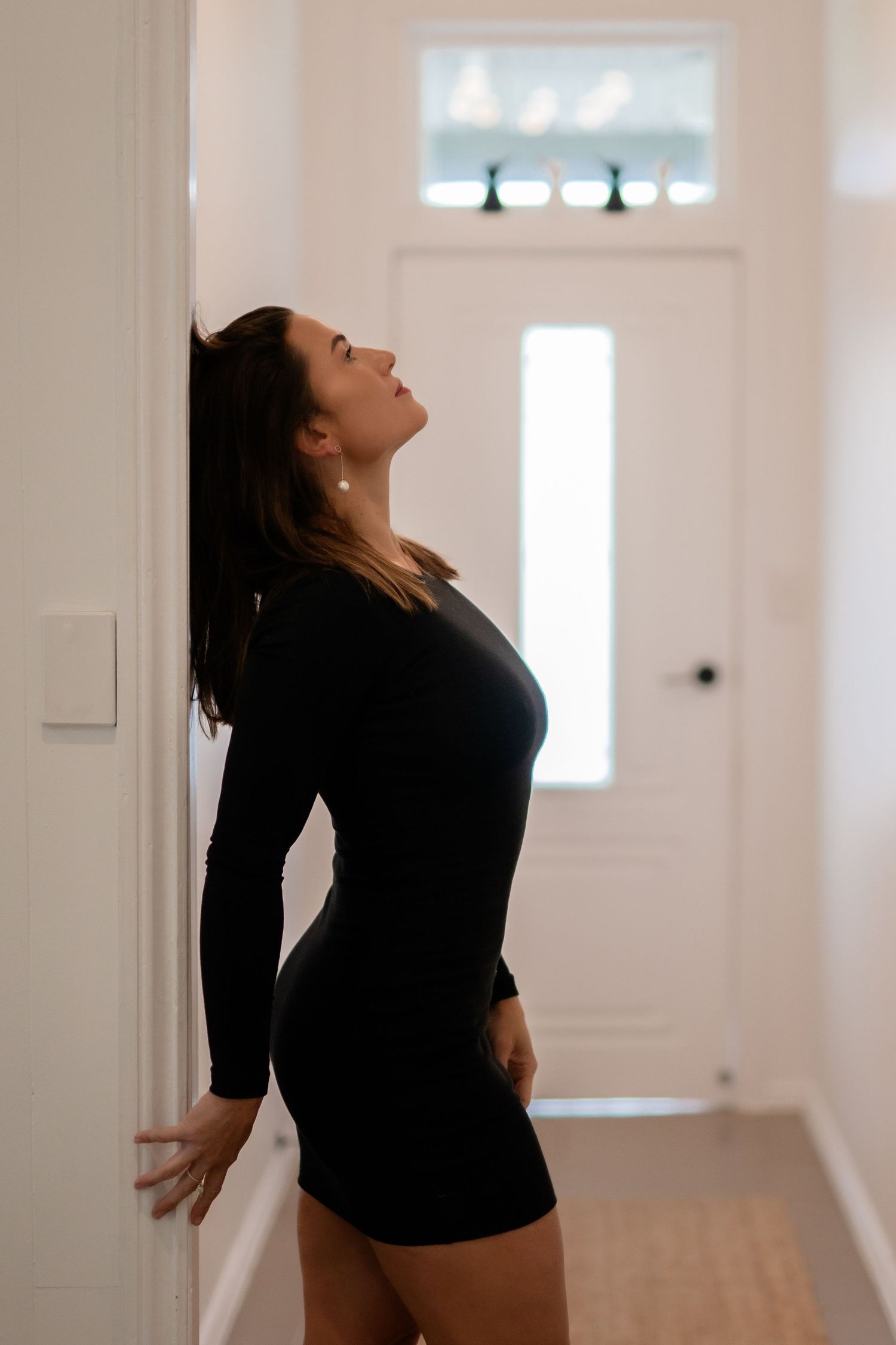 Woman in black mini dress leaning against white wall in hallway, looking up gracefully.