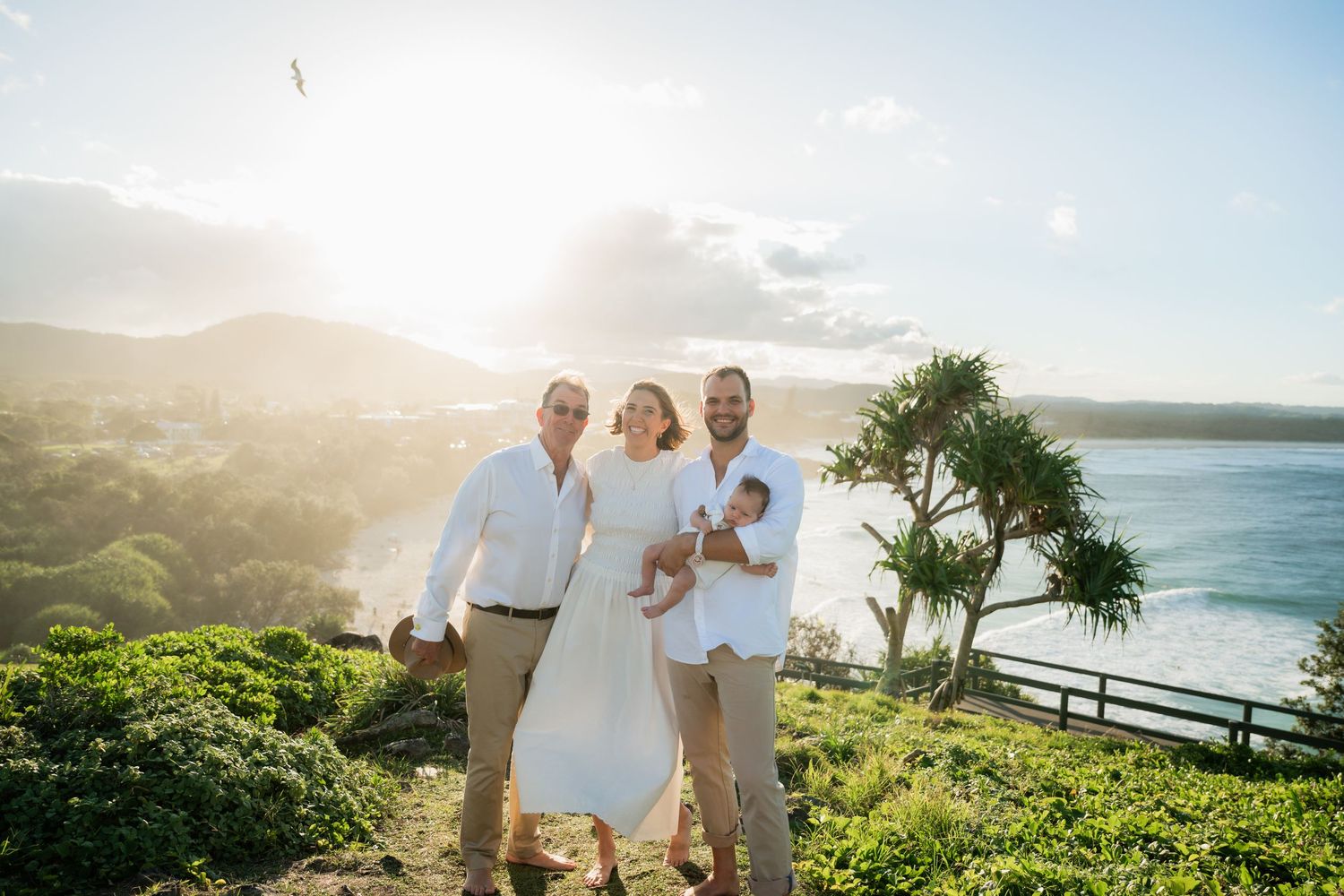 Happy family of three with a baby poses on a scenic coastal hilltop at golden sunset.