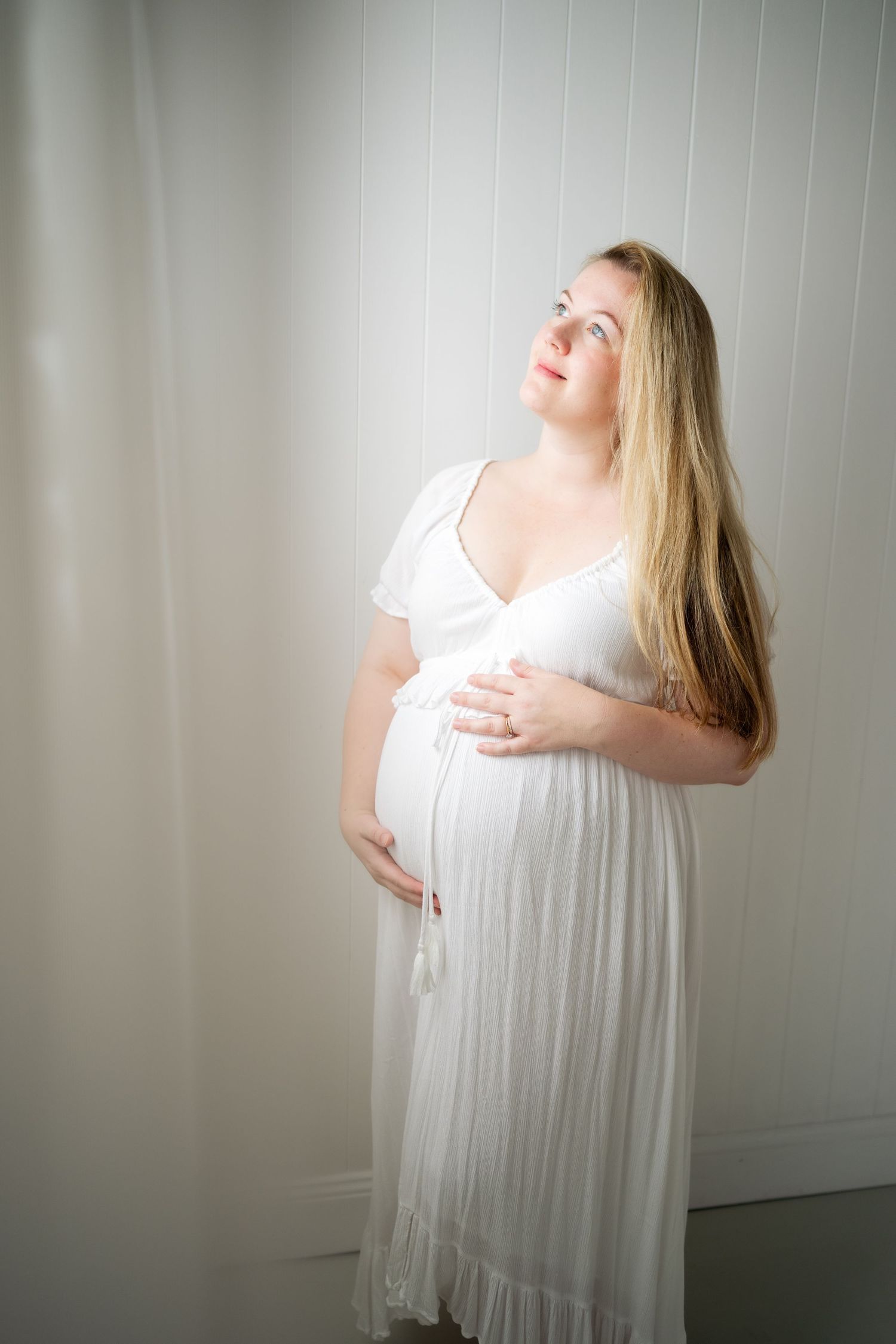 Pregnant woman in white dress cradling baby bump, looking up with a serene smile in a bright, airy room.