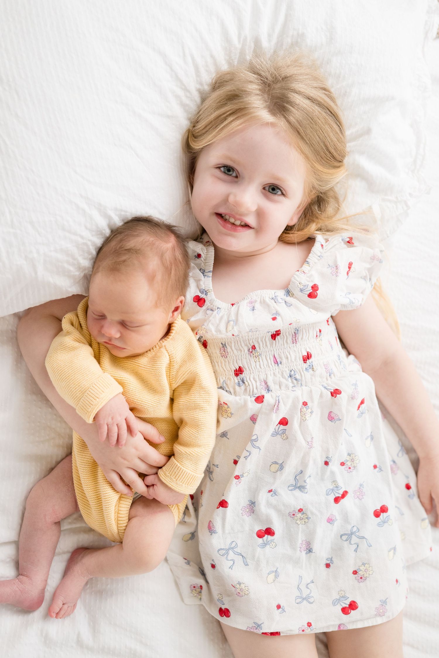 Young blonde girl in floral dress smiling beside newborn baby sibling lying on white bed.