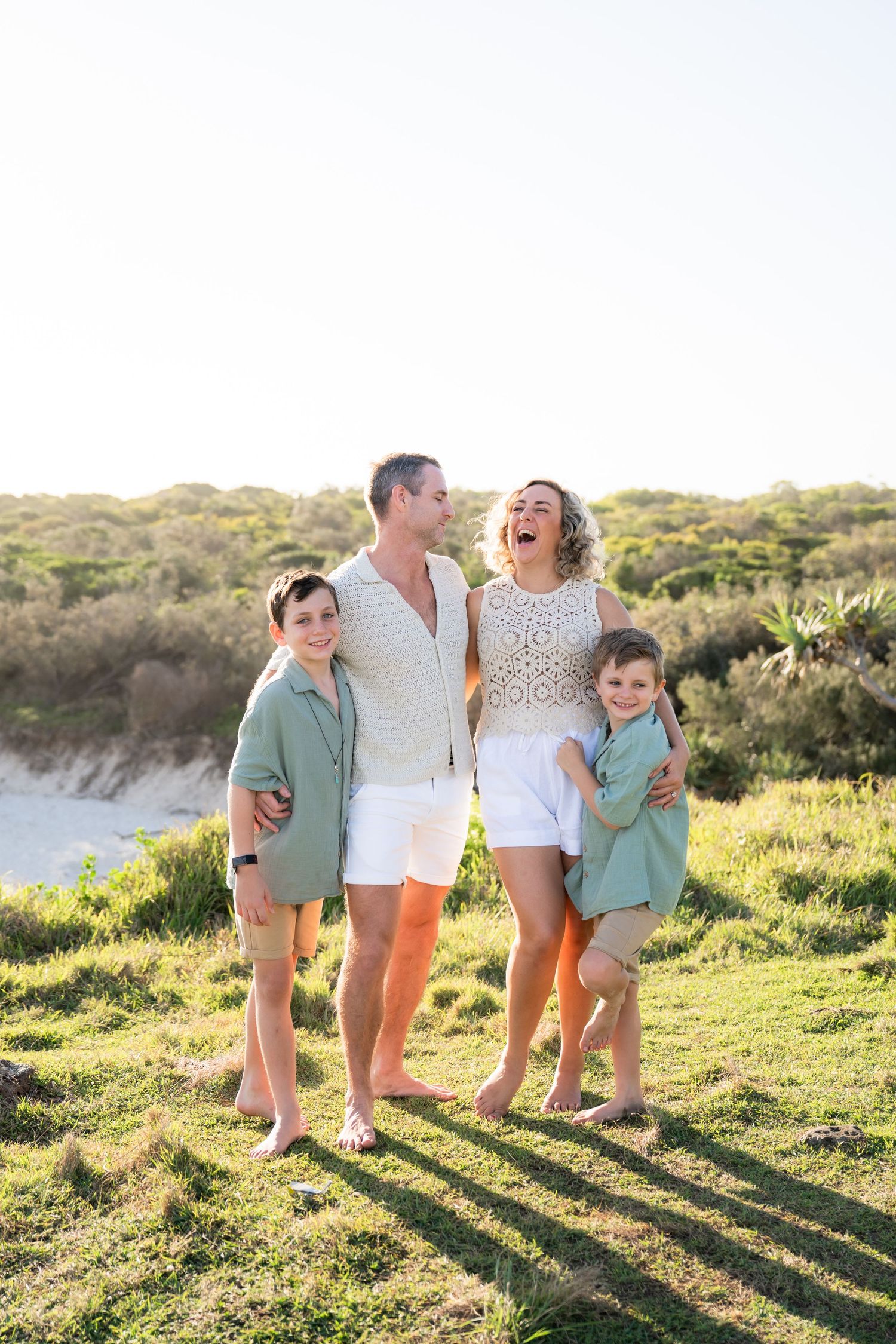 Happy family of four laughing and embracing outdoors on green grass near sandy dunes on a sunny day.