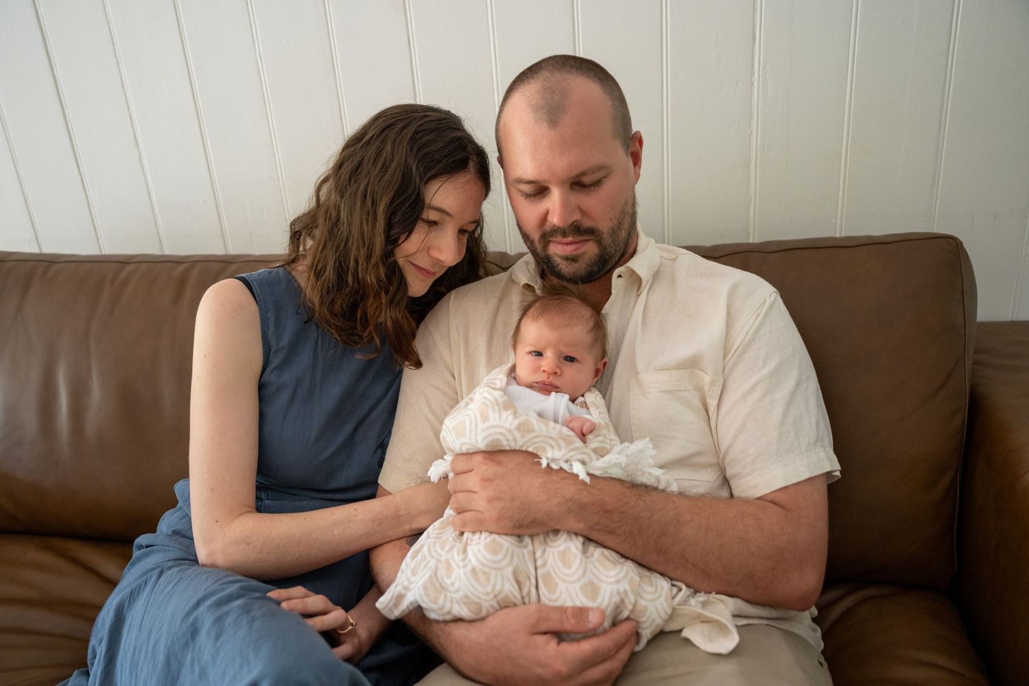 A couple sitting on a brown leather sofa tenderly holding their newborn baby dressed in white.