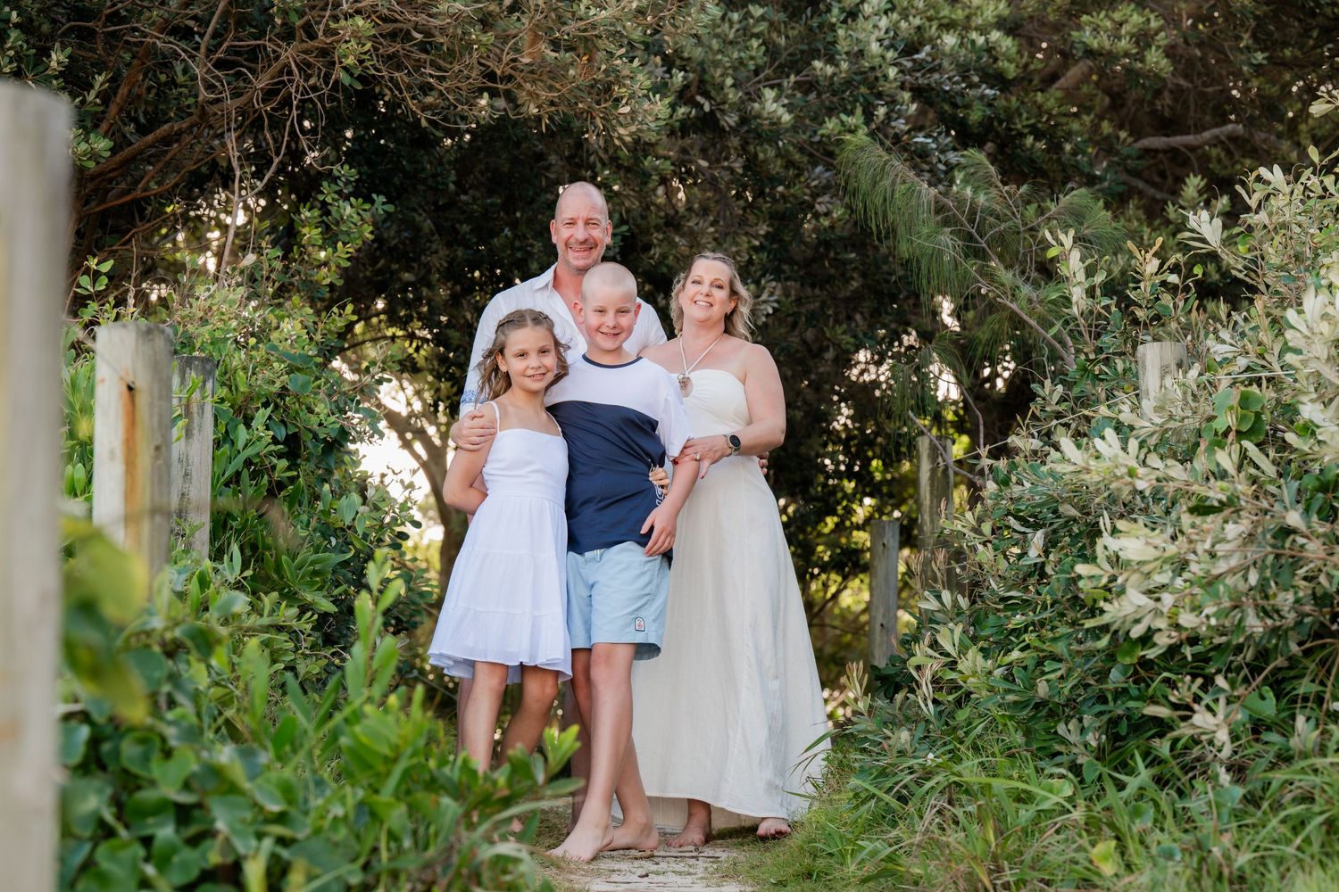 Family of four posing on a garden path surrounded by lush greenery, smiling warmly in casual summer attire.