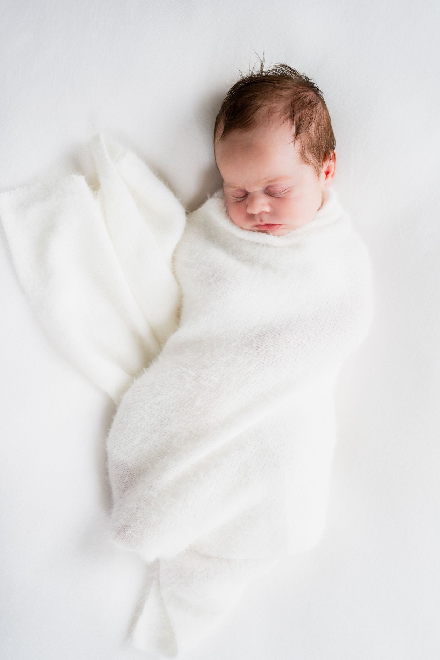 Peaceful newborn baby swaddled in a soft white blanket, sleeping on a white background.