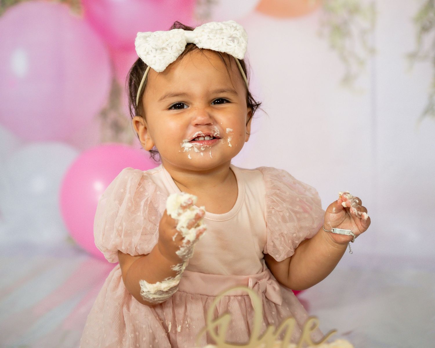 Baby in pink dress enjoys smashing and playing with birthday cake during first birthday cake smash photoshoot.
