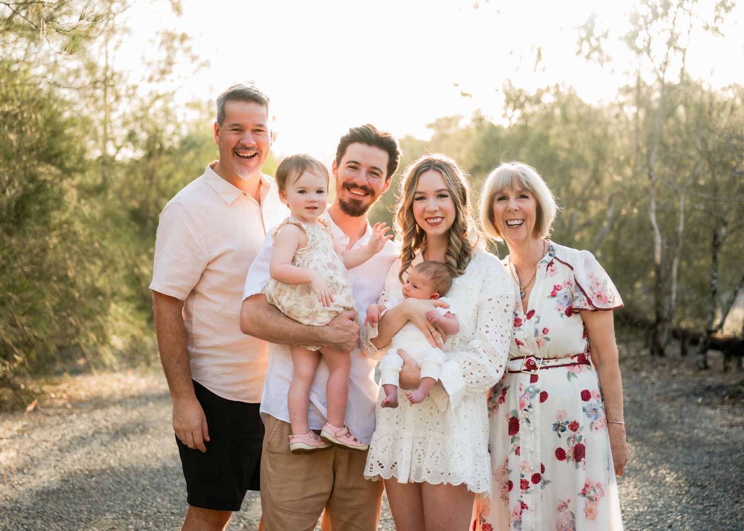 Six smiling family members pose outdoors on a gravel path surrounded by trees in warm golden light.