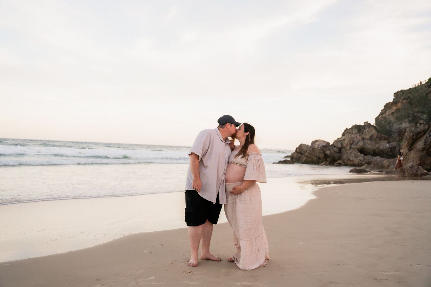 A romantic couple embraces on a scenic beach at sunset with rocky cliffs in the background.
