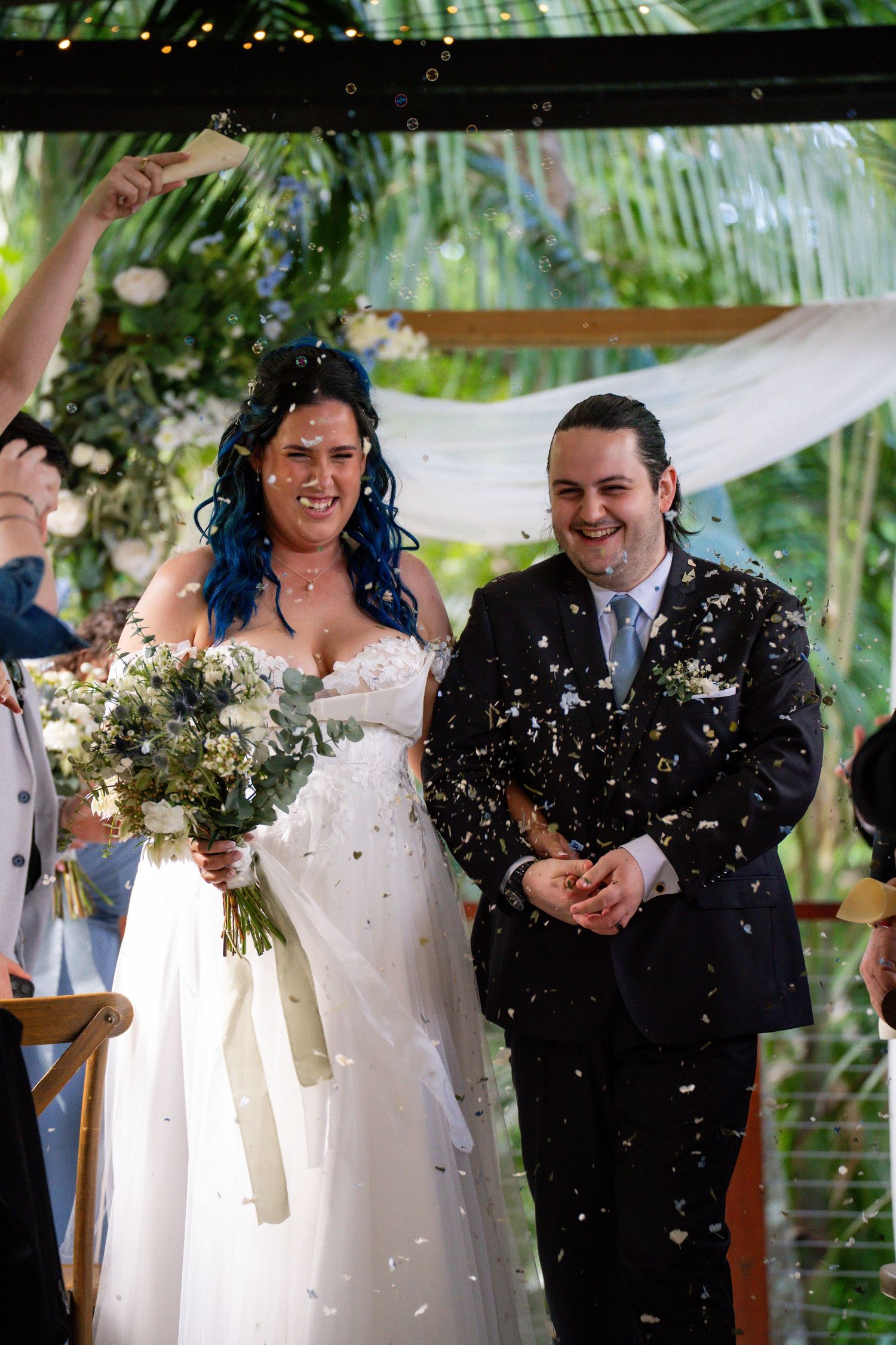 Newlywed couple laughs joyfully while being showered with confetti at an outdoor garden wedding ceremony.