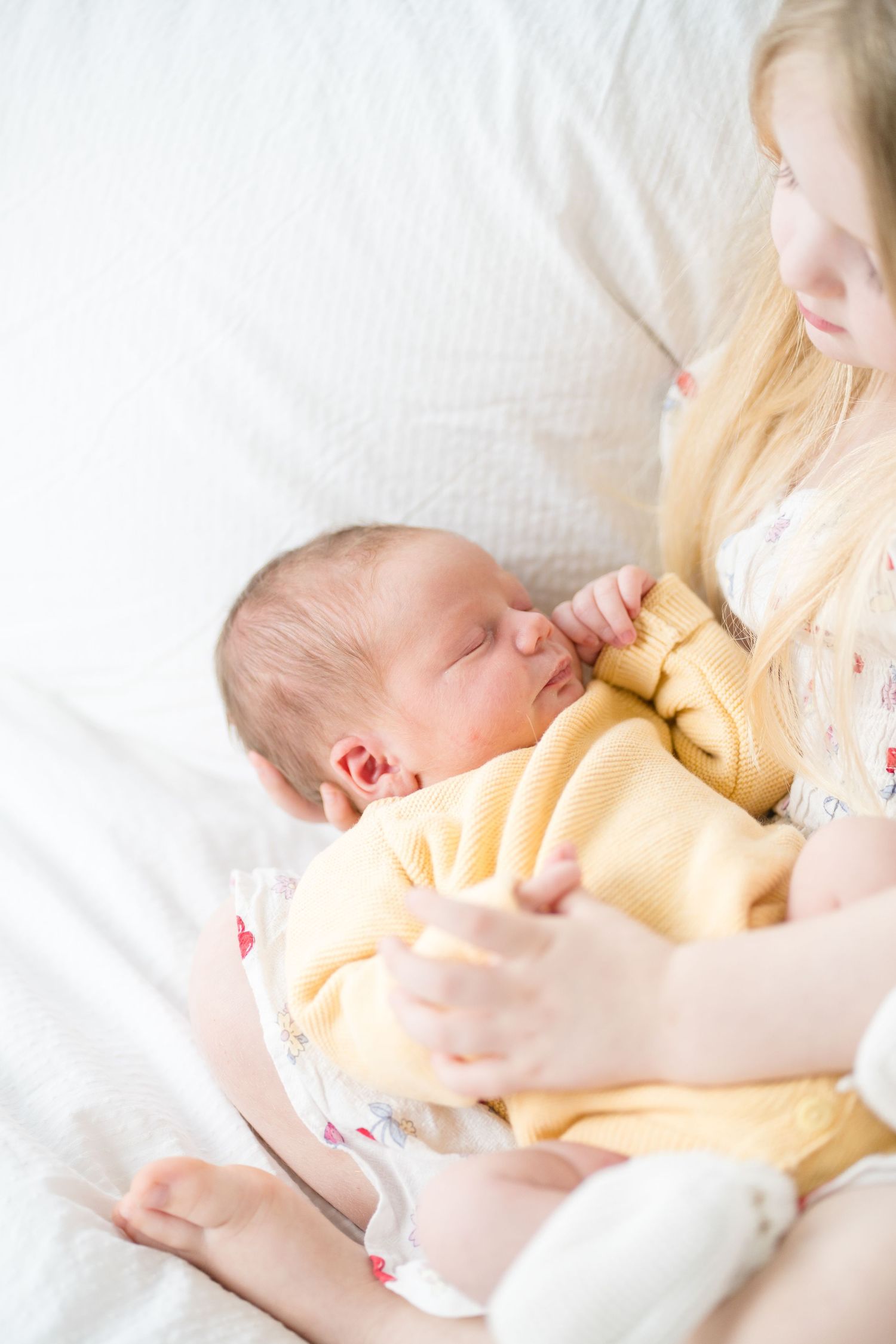 A young girl with blonde hair gently holds a sleeping newborn baby dressed in a yellow outfit on a white bed.