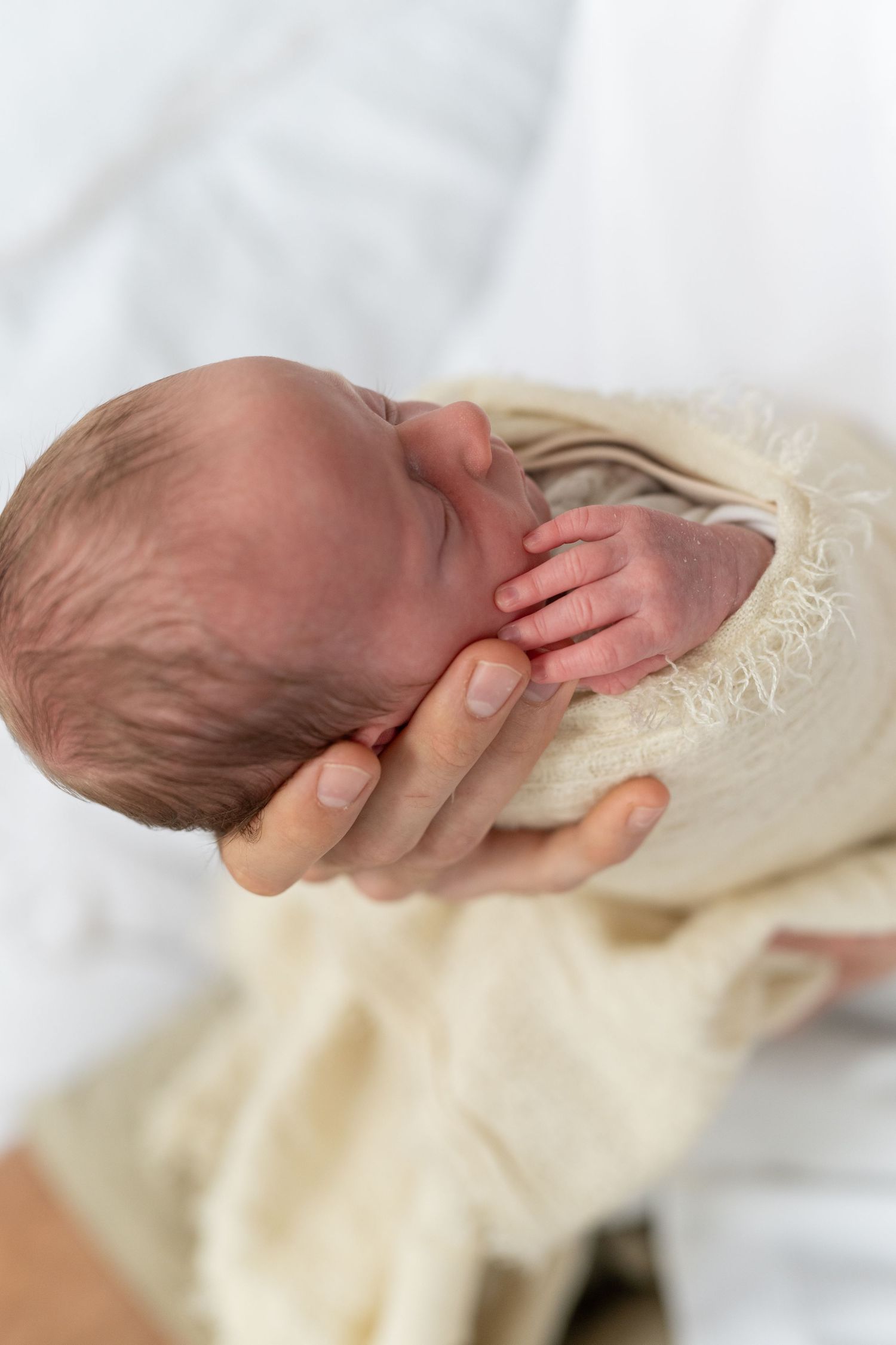 Newborn baby swaddled in cream wrap cradled gently in adult hands, tiny fingers visible against palm.