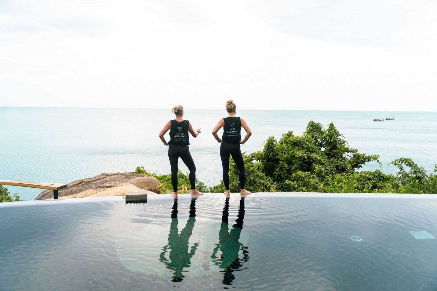 Two women in black activewear stand at infinity pool edge overlooking lush tropical ocean view.