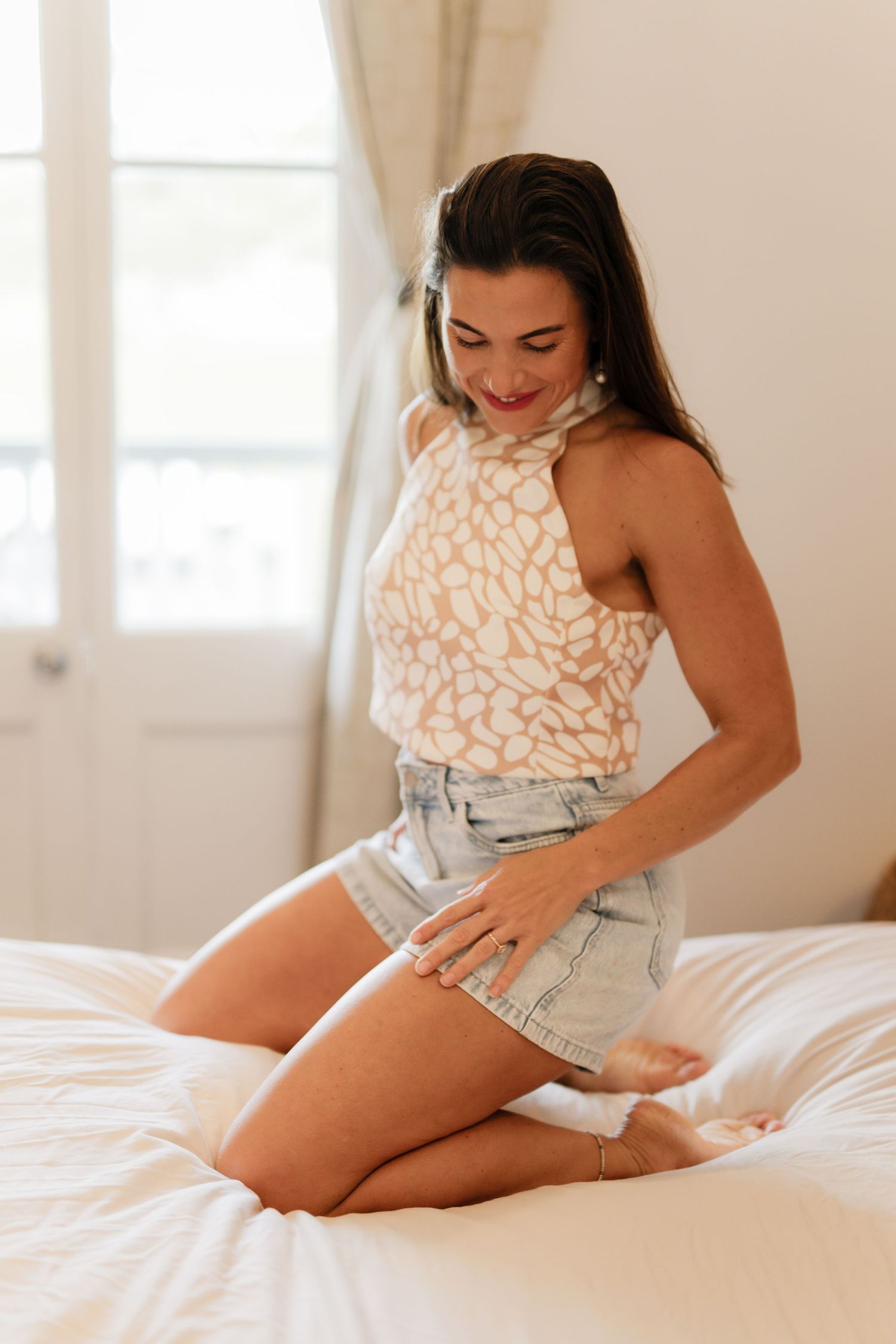 A young woman in a patterned halter top and denim shorts kneeling on a white bed, smiling downward.