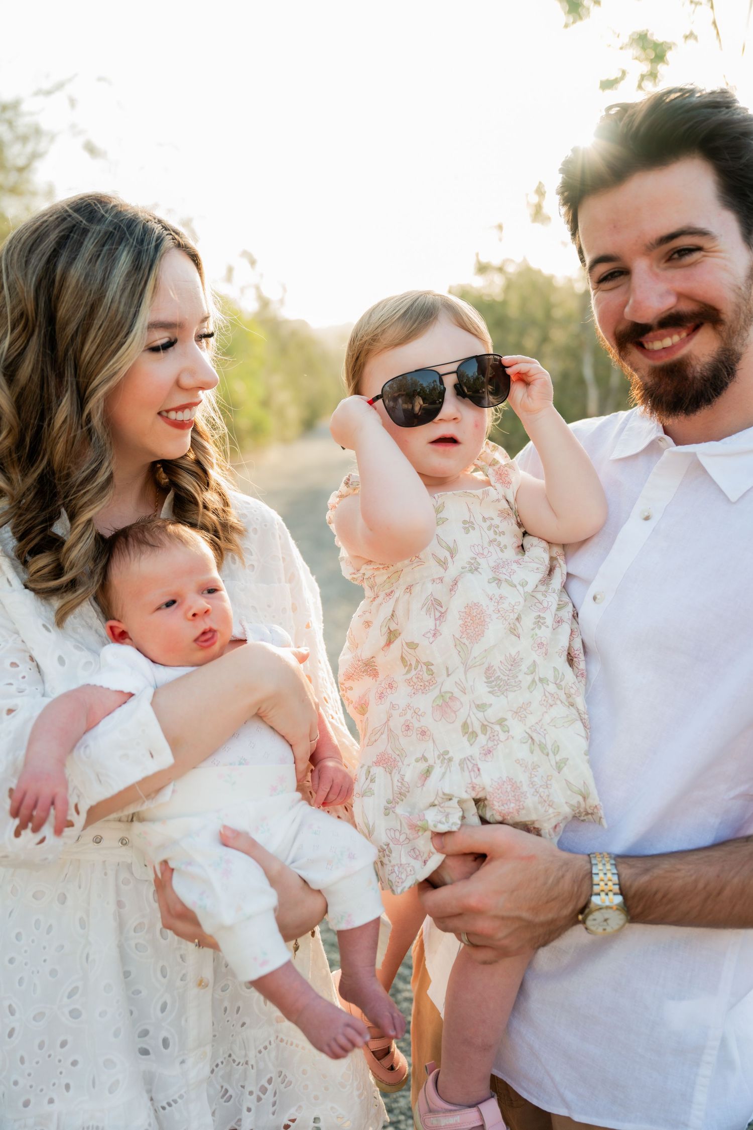 A smiling family with two young children outdoors, toddler wearing oversized sunglasses, warm golden hour lighting.