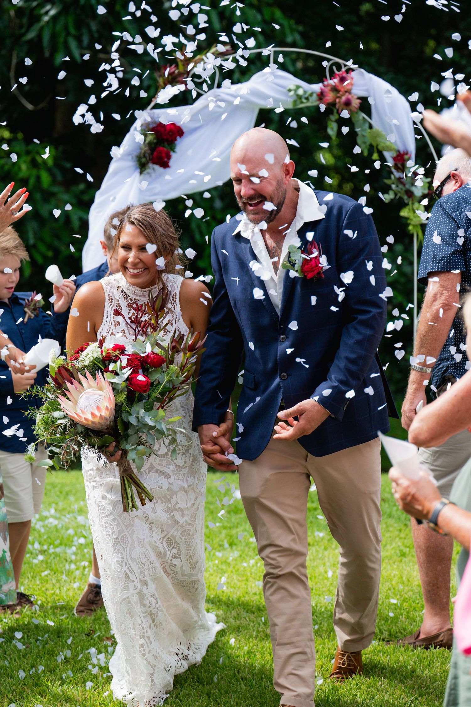 Bride and groom walk down the aisle as guests throw white confetti petals at outdoor wedding ceremony.