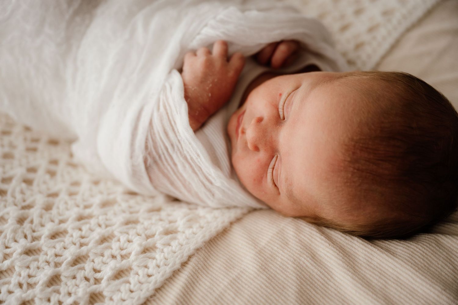 Peaceful newborn baby swaddled in white wrap sleeping on a soft cream crochet blanket.