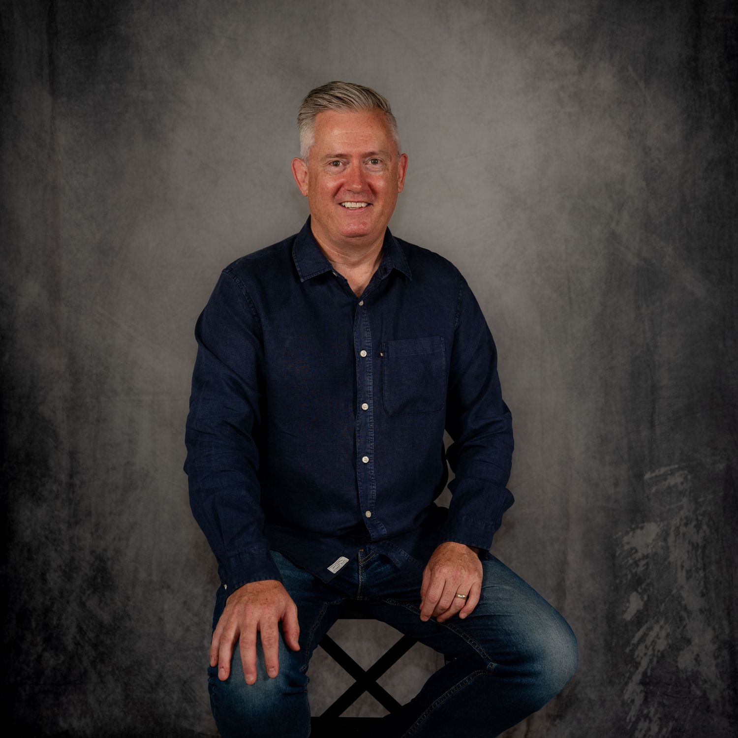 Professional studio portrait of someone in a dark blue button-down shirt against a textured gray background.