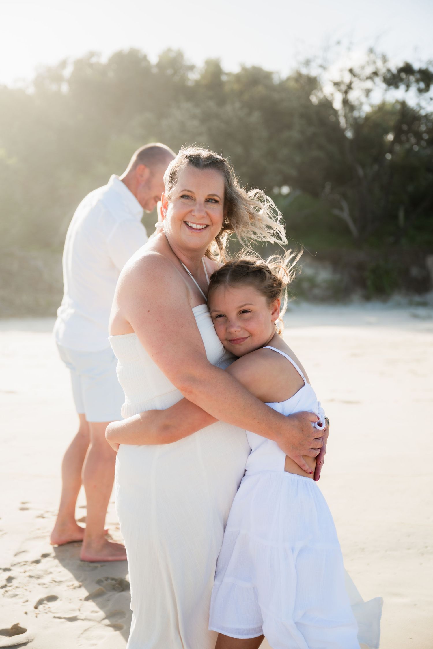 Woman and child hugging on sandy beach in white outfits with man standing behind them in golden sunlight.