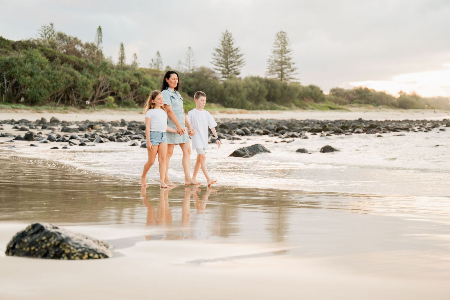 A mother and two children walk along a sandy beach with rocks and green trees in the background.