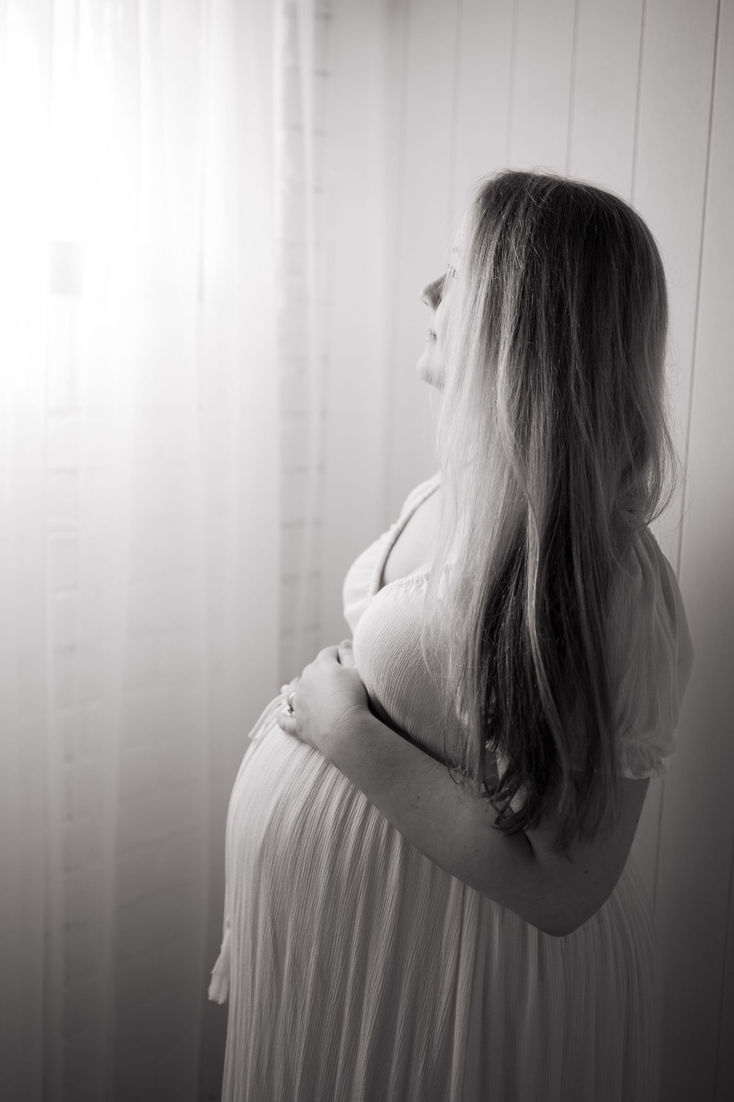 Black and white photo of a pregnant woman in a white dress cradling her baby bump while standing by a window.