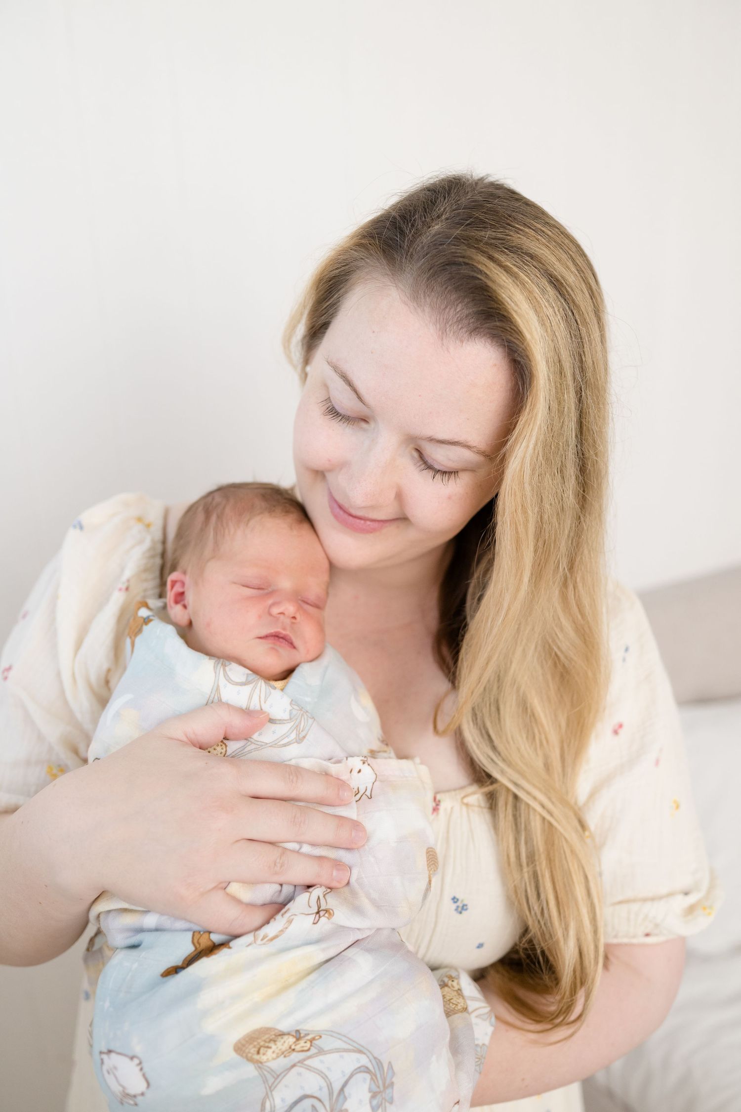 A smiling mother with long blonde hair tenderly holds her sleeping newborn baby wrapped in a soft swaddle blanket.