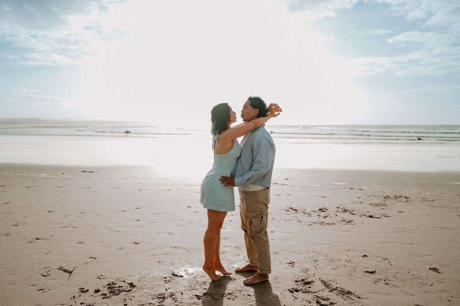 Couple embracing romantically on a sandy beach with ocean waves and bright sky in the background.