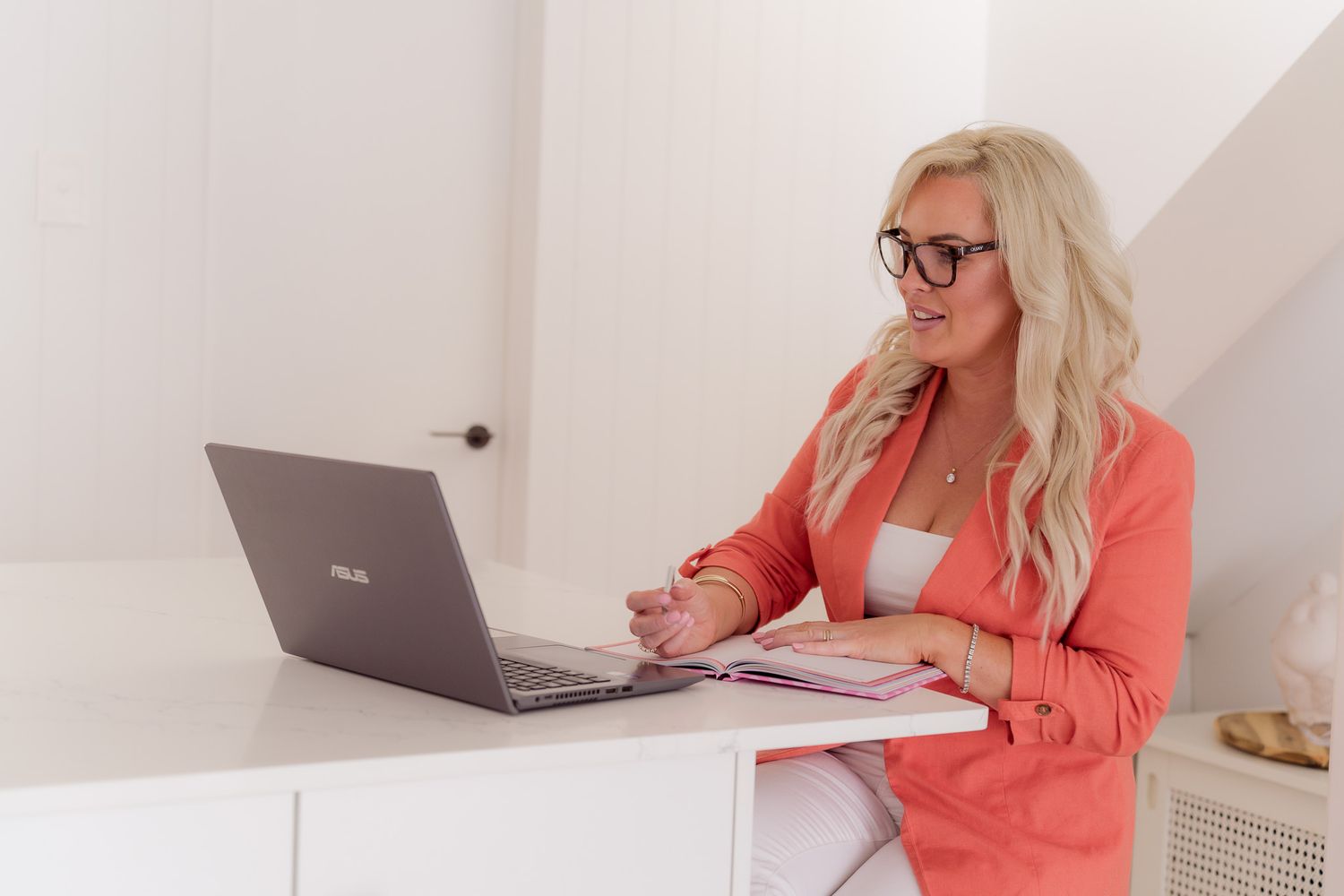 Professional working at a modern white desk with laptop and coral blazer in bright home office setting.