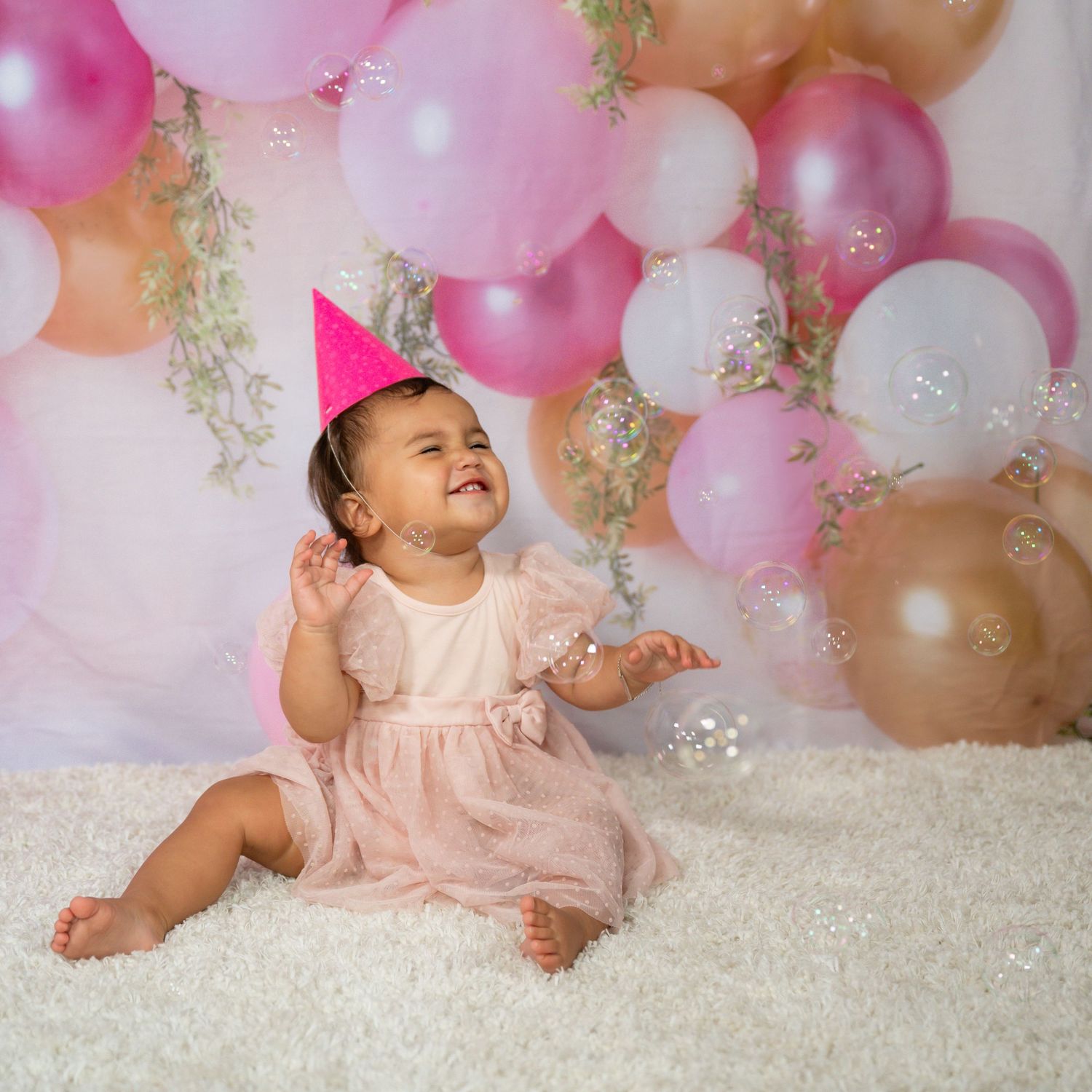 A smiling baby girl in a pink dress and party hat sits on white carpet surrounded by pink and gold balloons.
