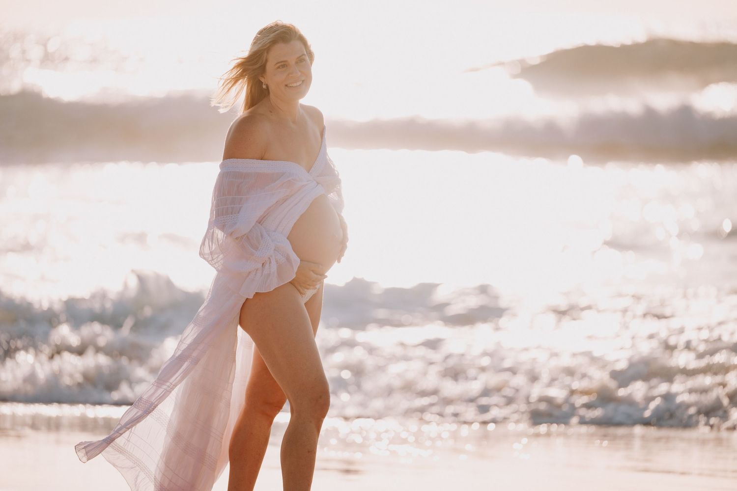 Pregnant woman in flowing white fabric poses on a sunlit beach with ocean waves in the background.