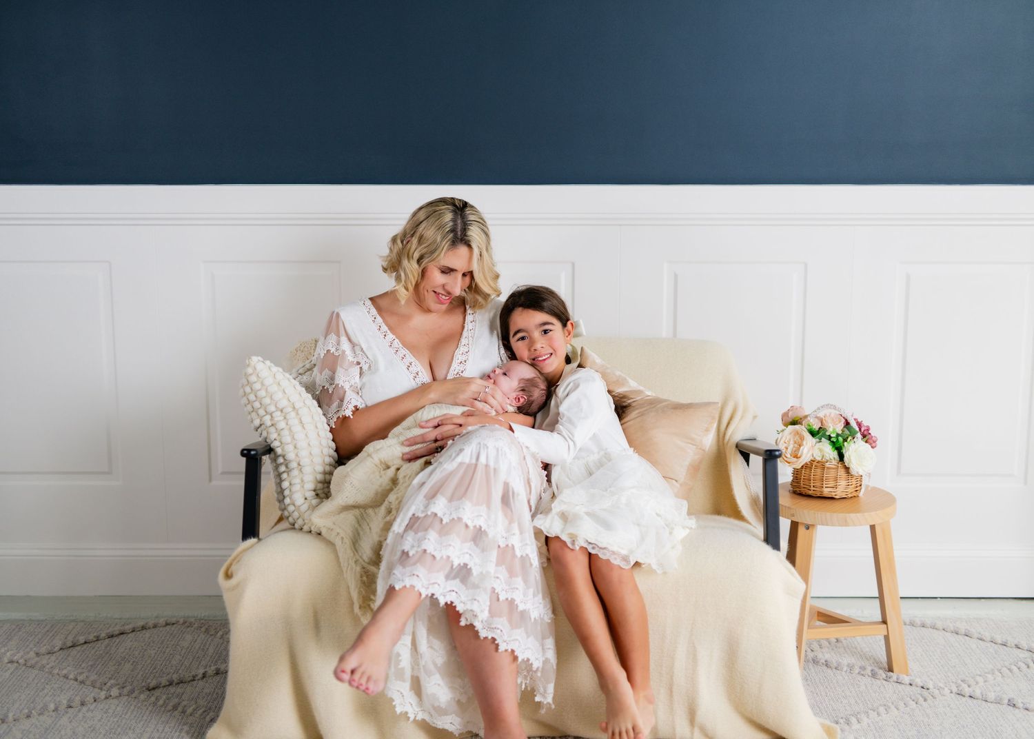 Mother and daughter in white lace dresses sitting together on a cream chair, lovingly holding a newborn baby.