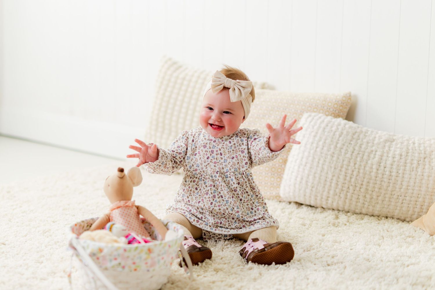 Happy baby girl in floral dress sitting on white fluffy rug, laughing with arms raised, surrounded by toys.