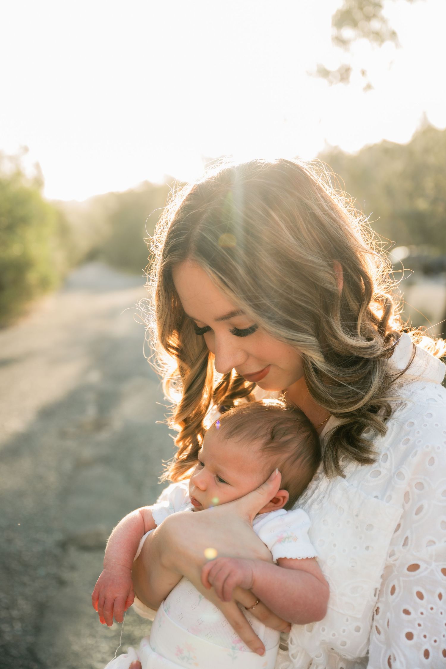 A mother tenderly holds her newborn baby in golden sunset light outdoors, both dressed in white.