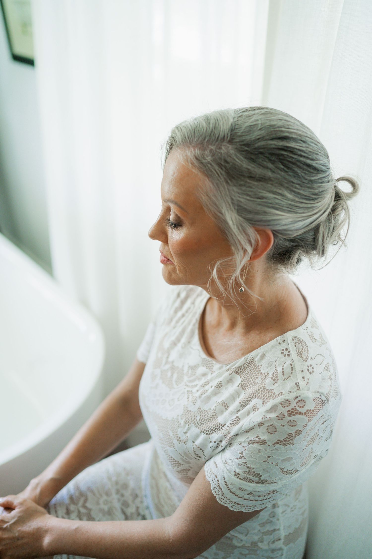 Elegant bride with silver updo in white lace dress sitting by a window in soft natural light.
