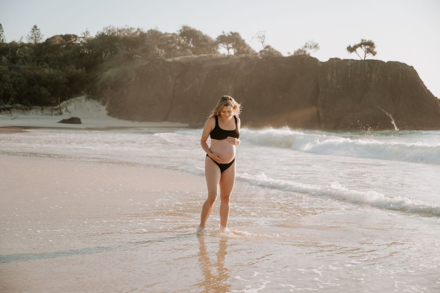 Pregnant woman in black bikini standing on a sunny beach with waves and rocky cliffs in the background.