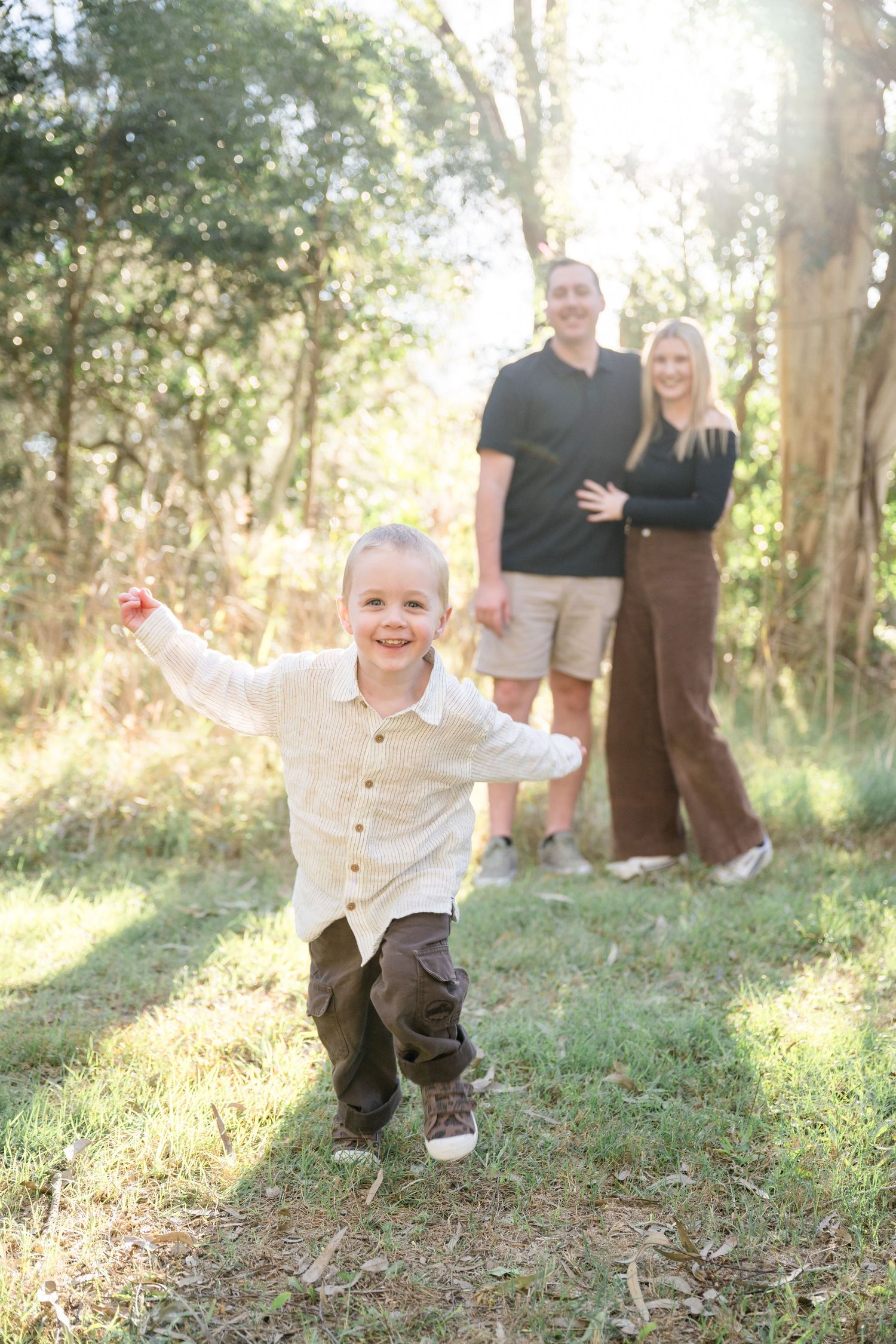 A joyful toddler runs toward the camera while his parents stand together in a sunlit forest clearing.