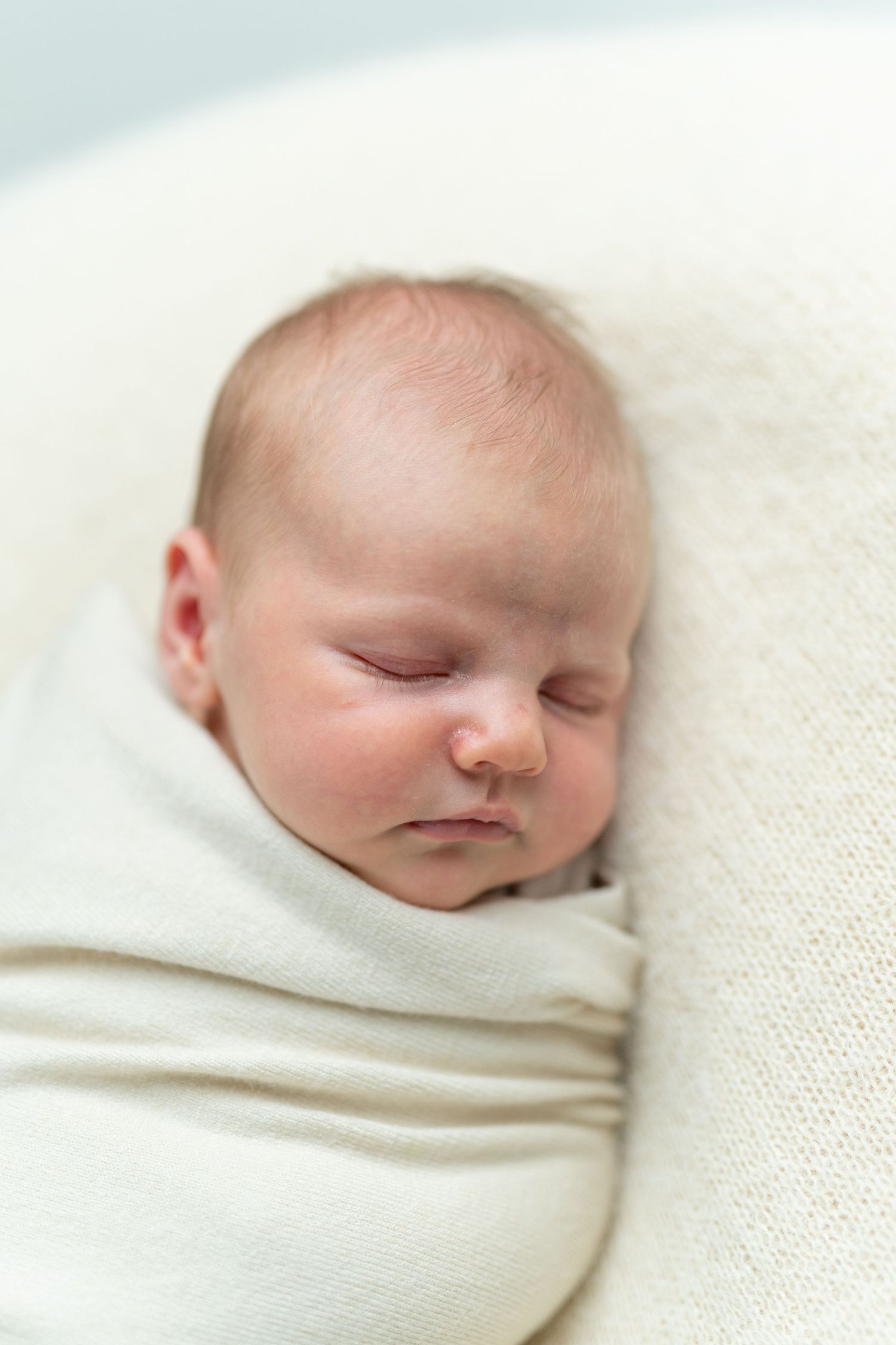 Peaceful newborn baby swaddled in a soft cream wrap, sleeping soundly in a white knit bowl.
