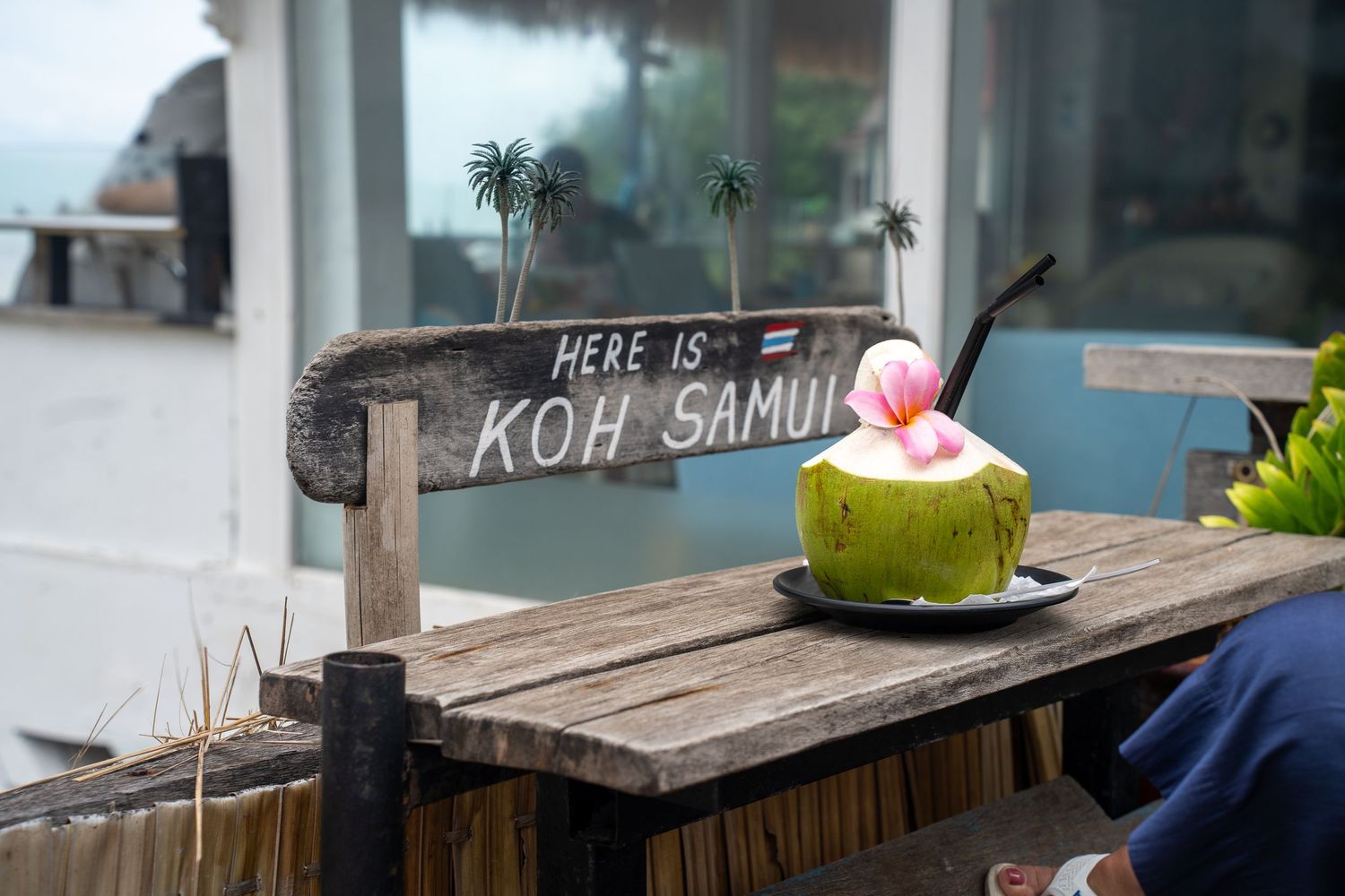 Fresh green coconut drink on wooden table with Koh Samui sign at tropical beach cafe in Thailand.
