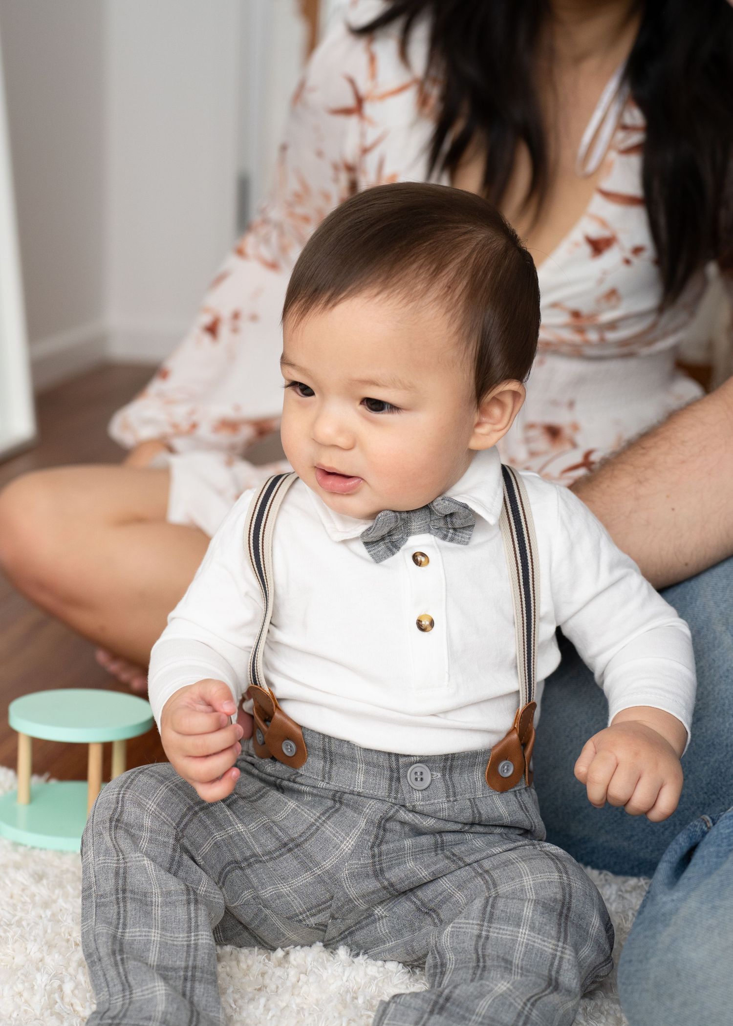 Adorable baby boy in white onesie with bow tie, suspenders, and plaid pants sits on floor with mother behind him.